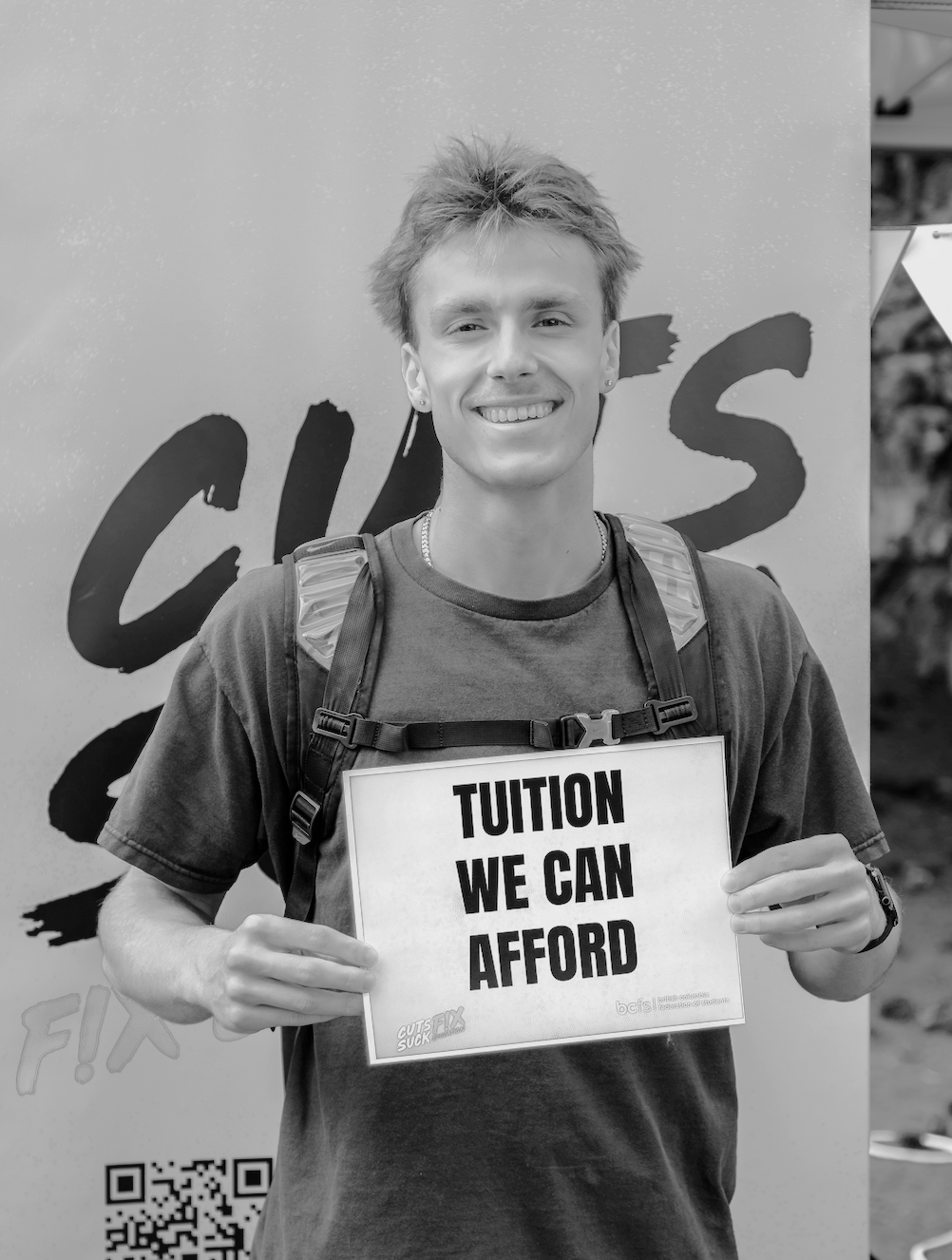 A young man with a backpack smiling and holding a sign that reads "Tuition We Can Afford" at an outdoor event.