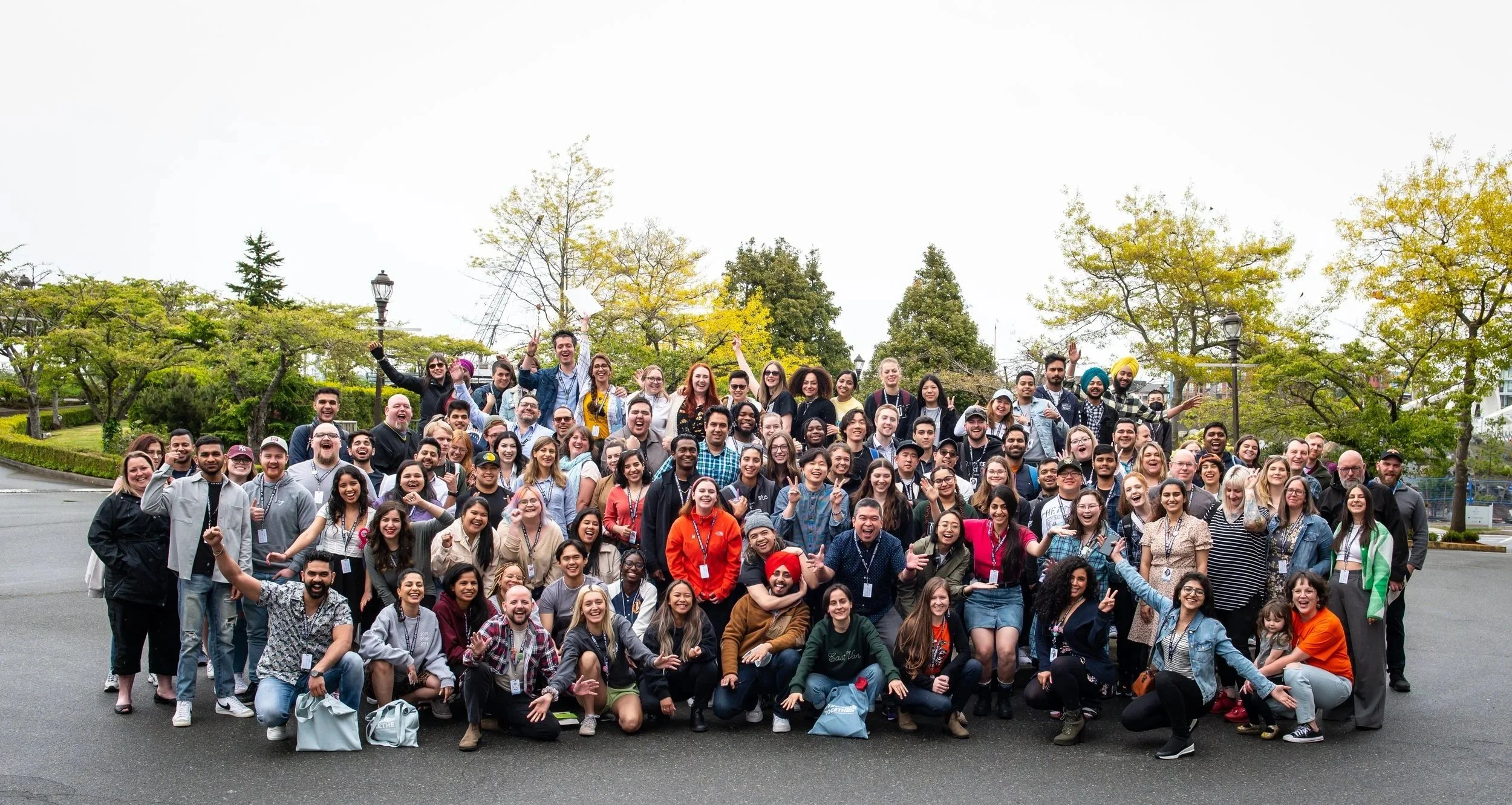 A large, diverse group of people standing outdoors in a park with trees and sky in the background, smiling and celebrating.