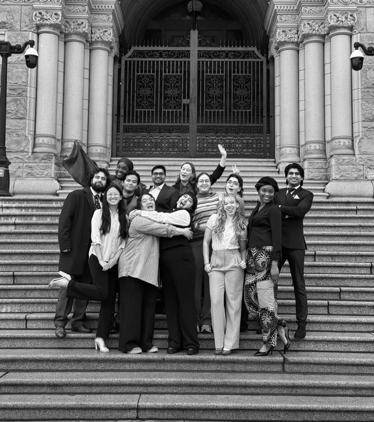 Group of diverse young adults celebrating on the steps of a historic building, smiling, hugging, and posing for a photo, with ornate architecture and gates in the background.