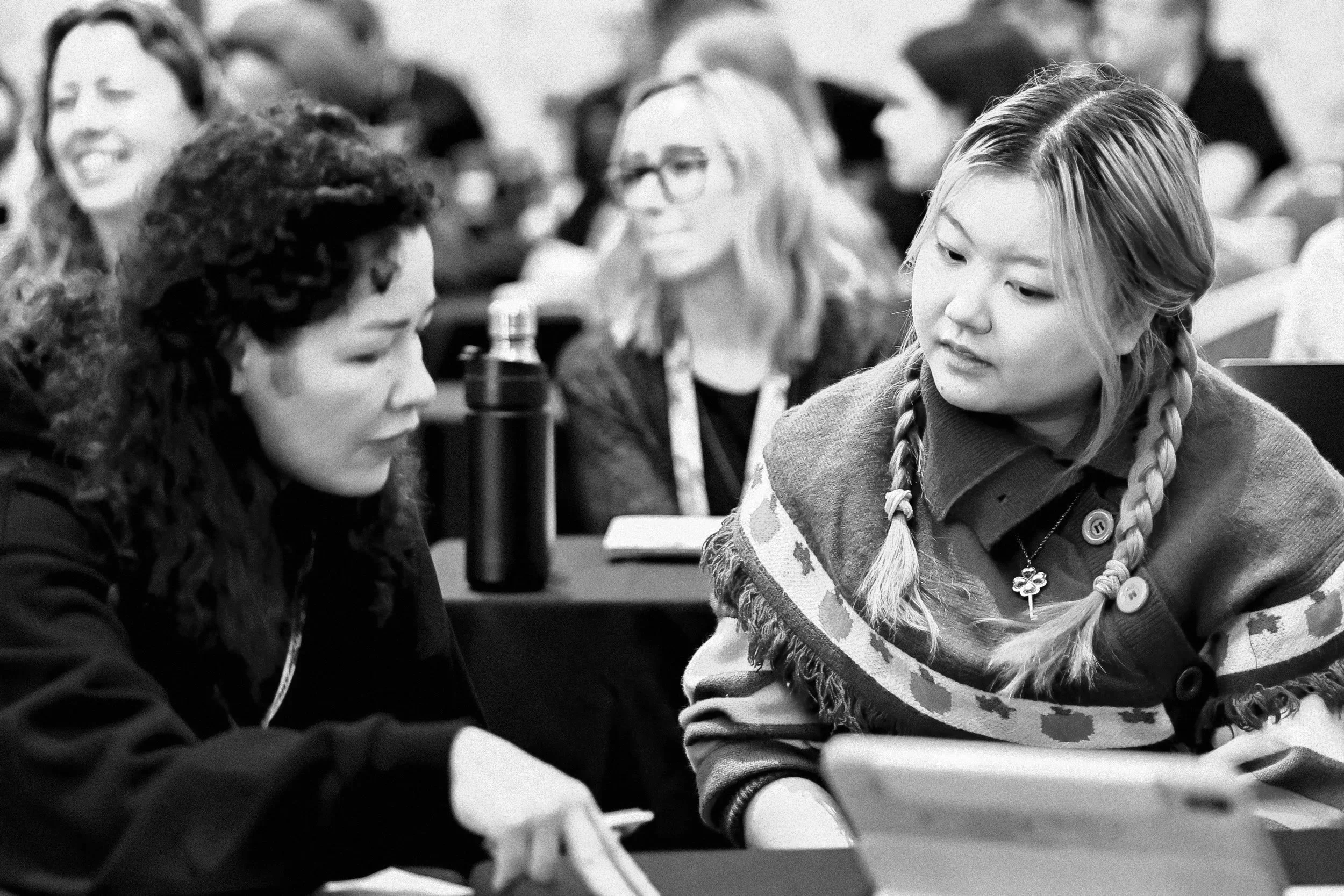 Two young women with braided hair and casual clothing are sitting together at a table, engaging in a conversation during a gathering or event, with other people blurred in the background.