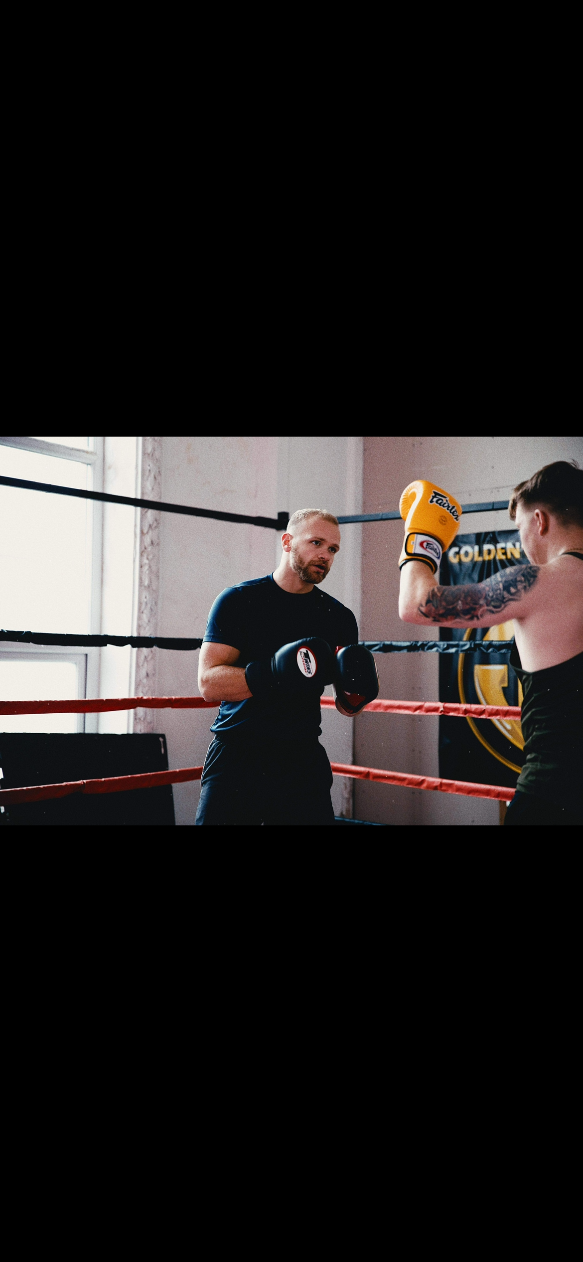 Two people training in a boxing gym. One person with a beard, wearing a black T-shirt and boxing gloves, is standing in front of another person with short hair, tattoos on their arm, and yellow boxing gloves. They are inside a boxing ring with ropes and a window in the background.