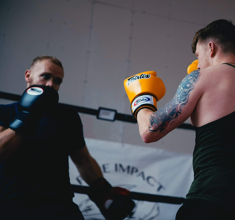 A boxing match in progress with one man throwing a punch and the other man blocking, both wearing boxing gloves in a gym setting.