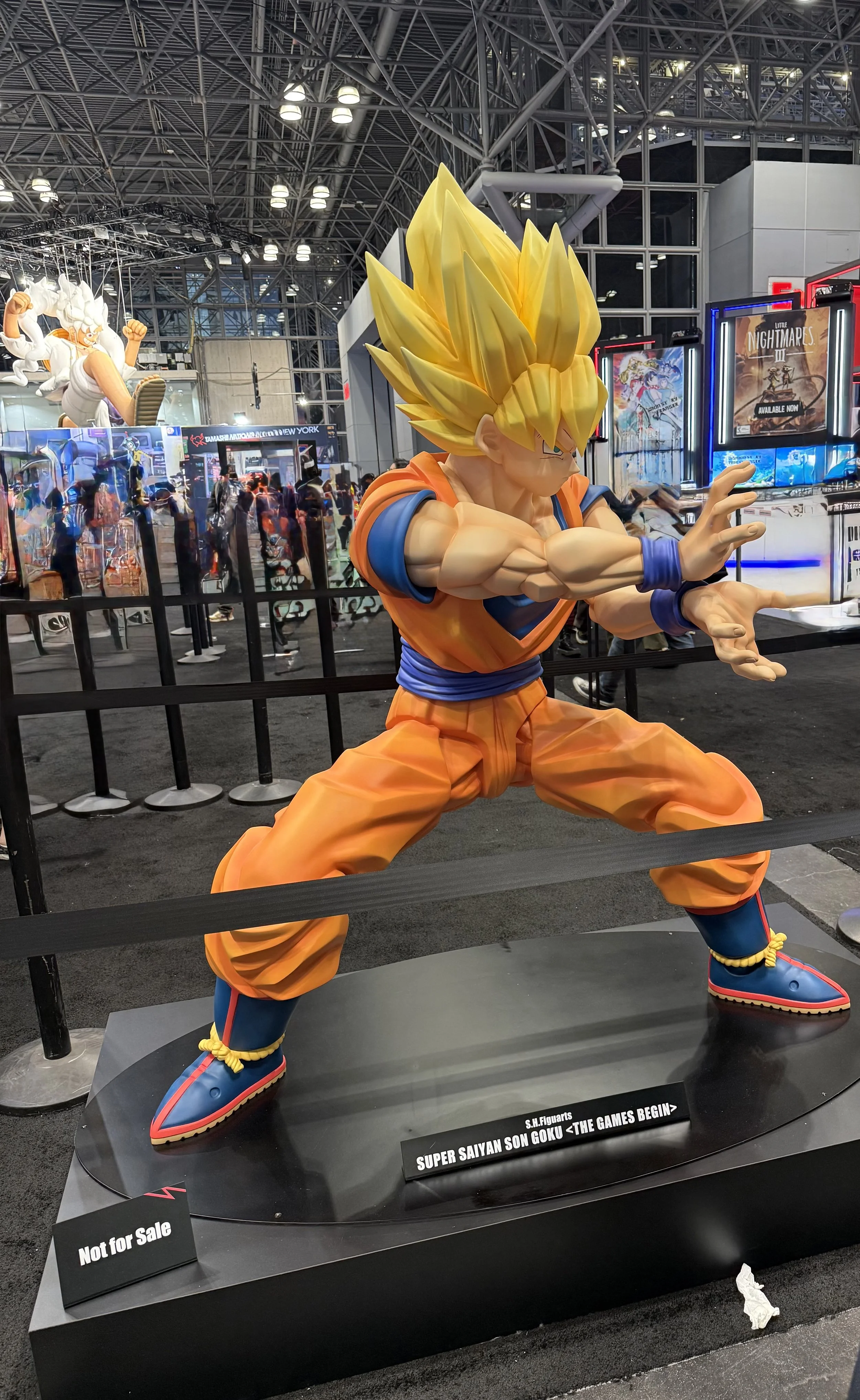 Life-sized model of Goku from Dragon Ball Z in a martial arts stance at a convention, with a sign beneath reading 'Super Saiyan Son Goku <The Games Begin>' and a smaller sign saying 'Not for Sale' in front.