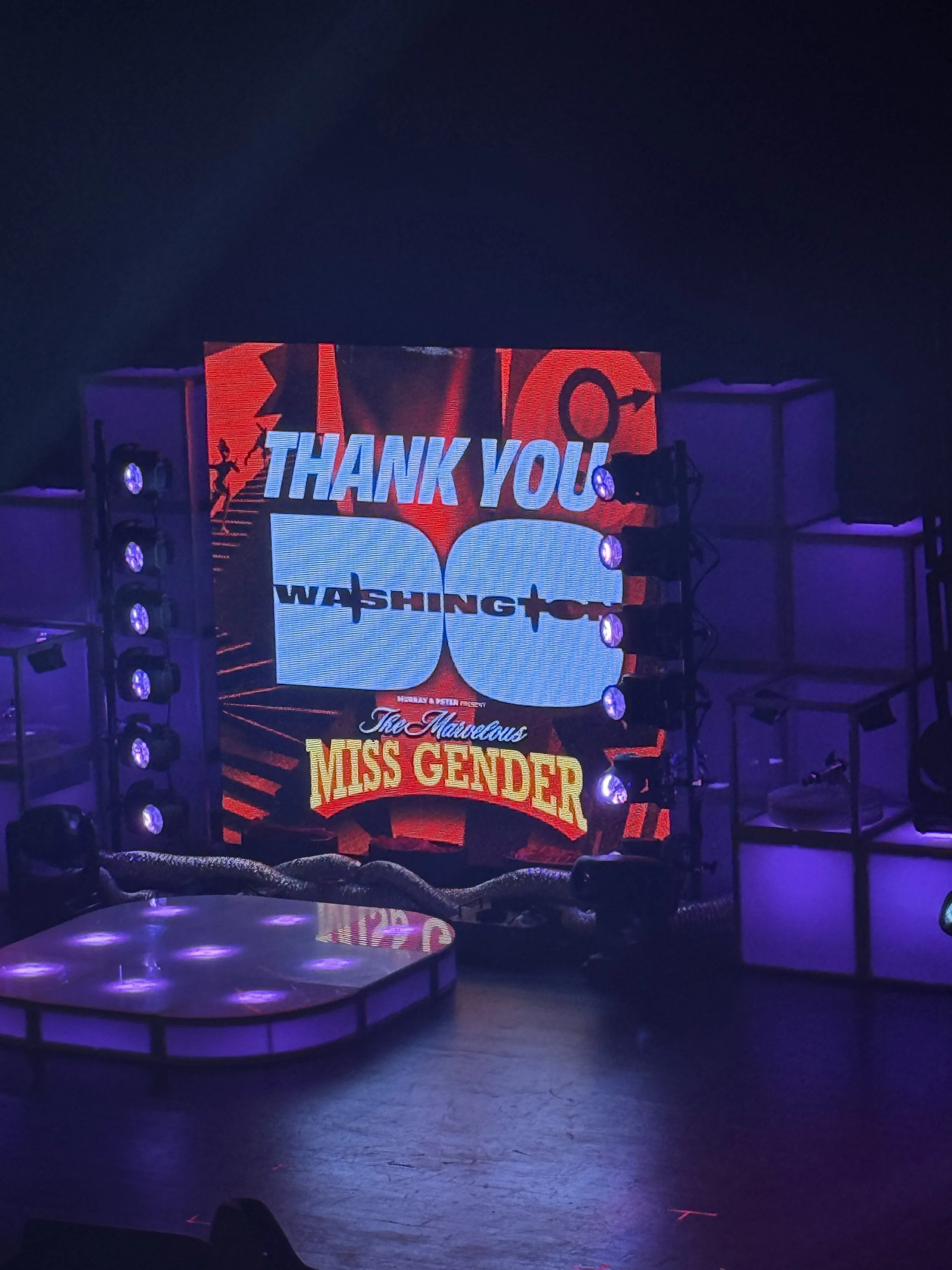 Stage with a large screen displaying a thank you message and visuals from the Miss Gender event, illuminated by purple and pink stage lights.