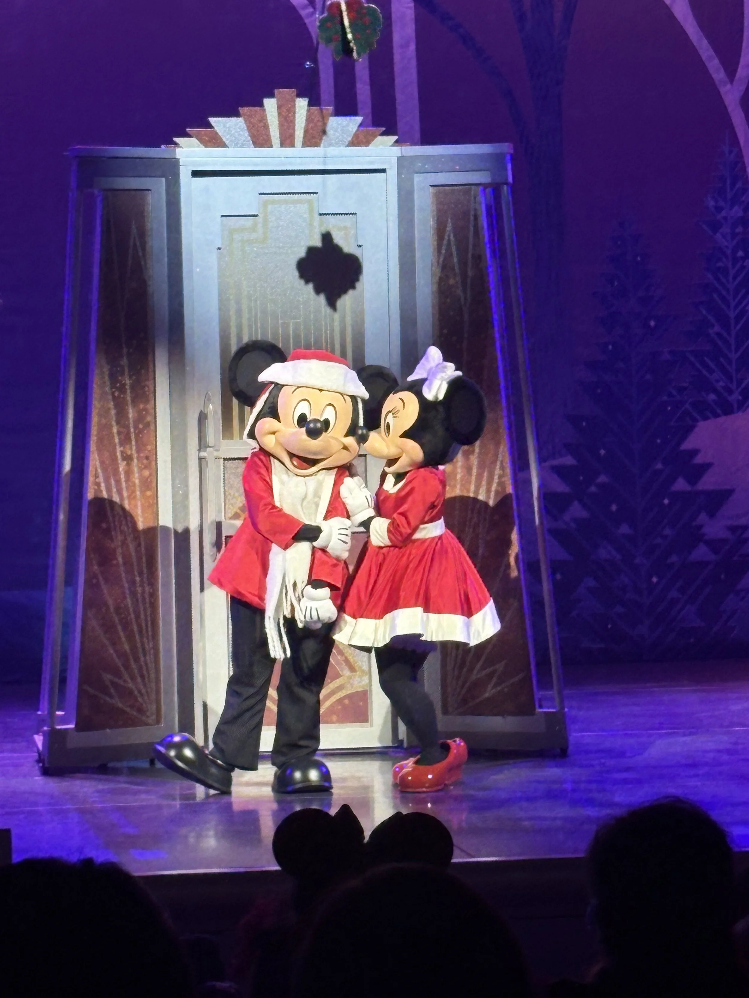 Mickey Mouse and Minnie Mouse dressed in Christmas outfits on stage, with a theatrical set background.