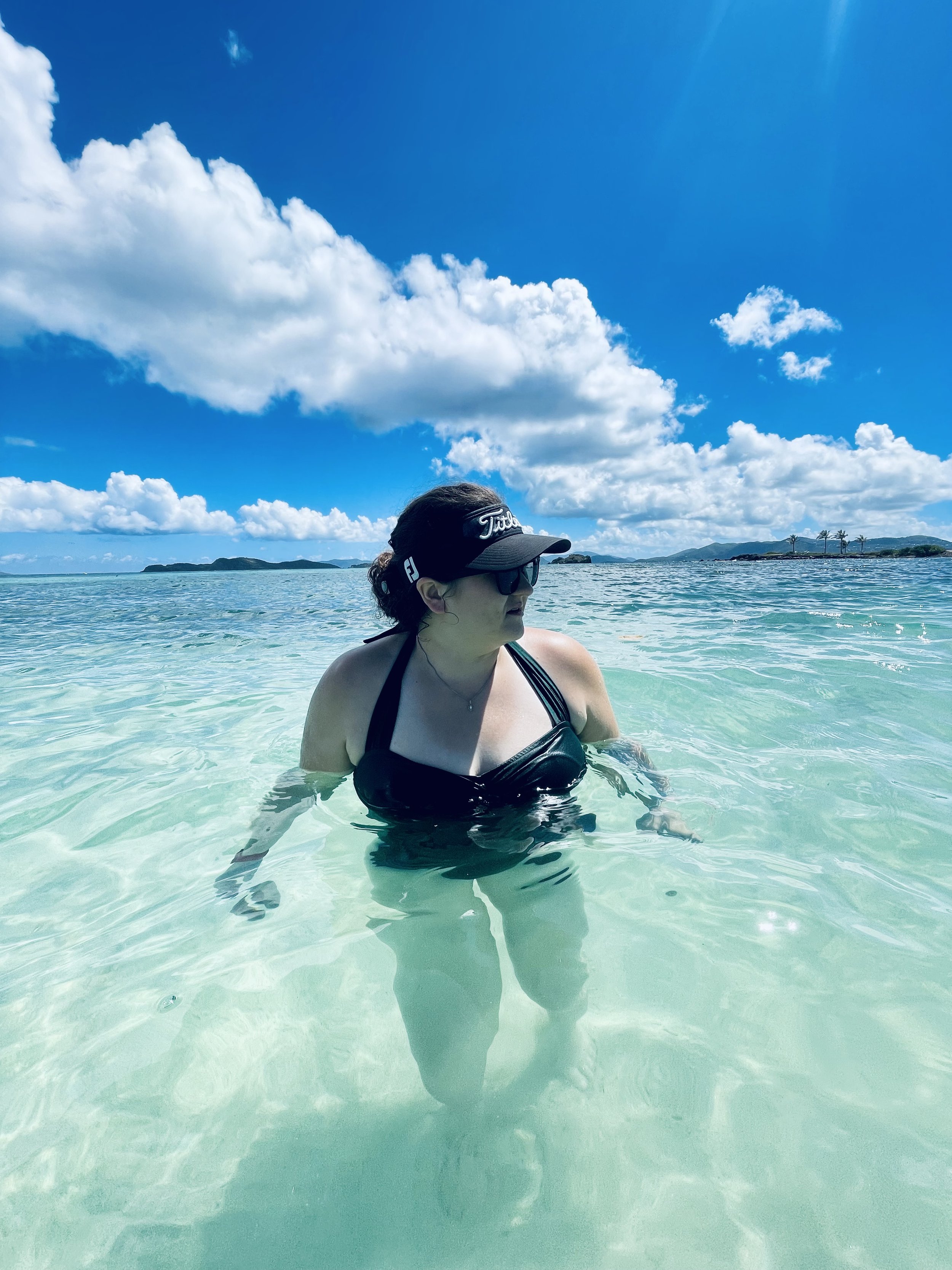 A woman in a black swimsuit, wearing sunglasses and a golf cap, wades in clear turquoise water with an island and palm trees in the background under a sunny sky with scattered clouds.