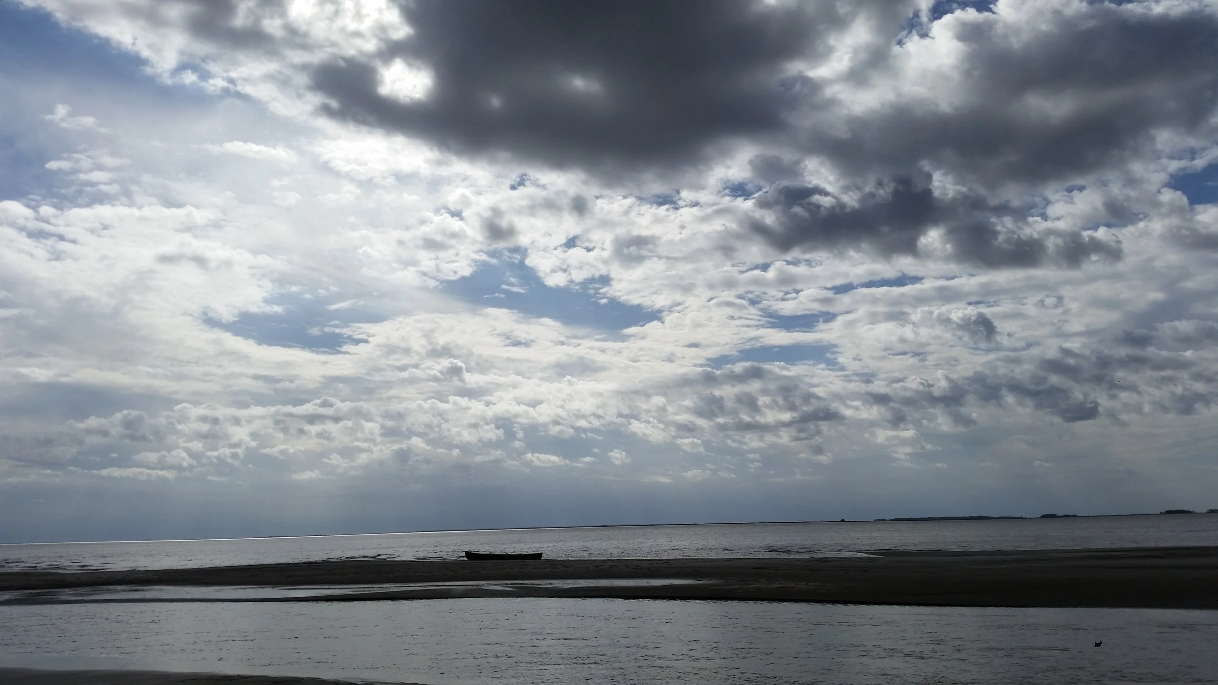A large body of water with a boat resting on the shore, under a cloudy sky with varying shades of gray and some patches of light, near a distant shoreline.