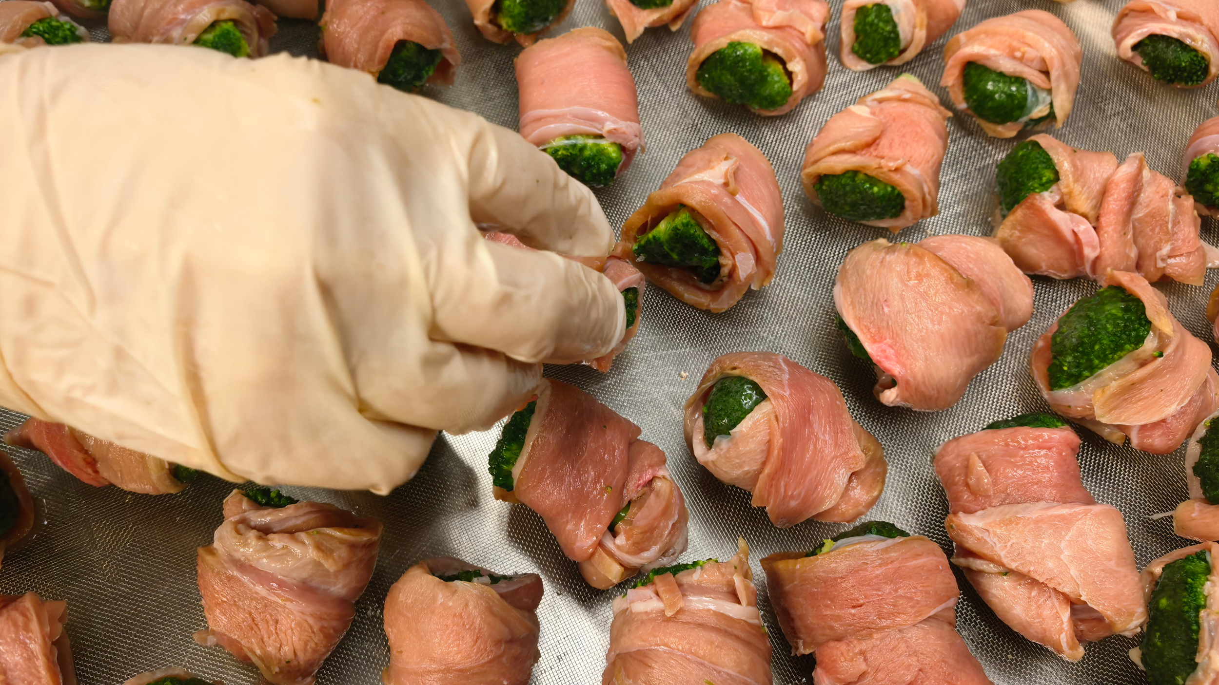 People preparing bacon-wrapped stuffed jalapeño peppers on a baking sheet.