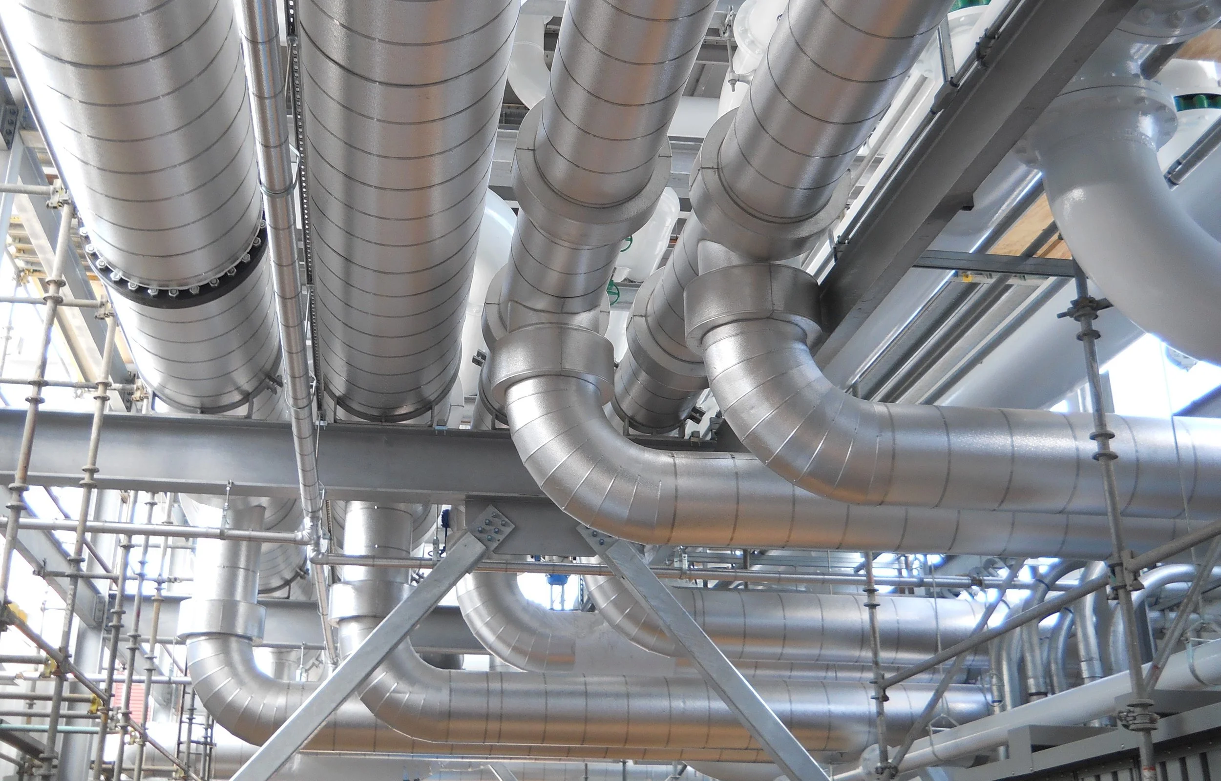 Multiple large metallic industrial pipes running parallel in an industrial facility.
