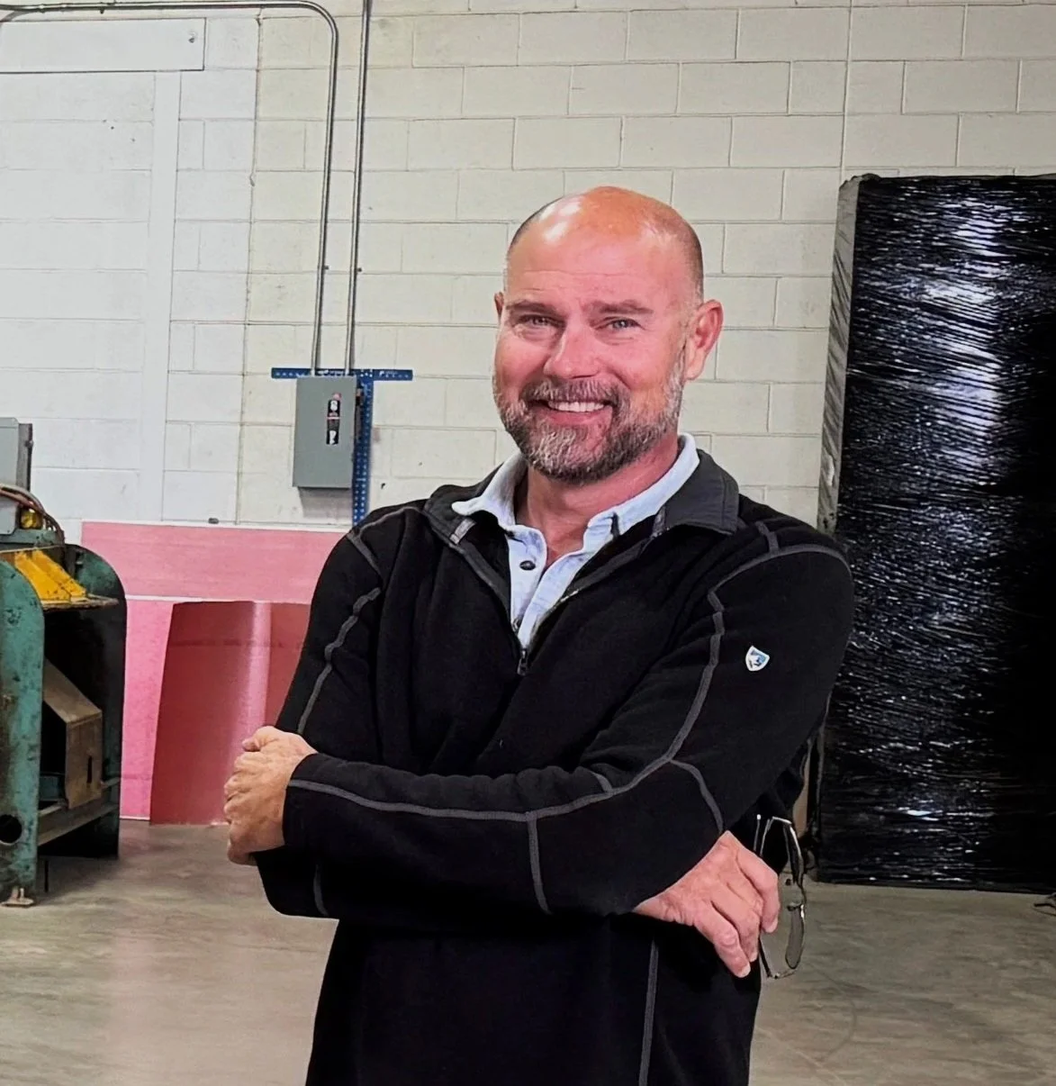 A smiling middle-aged man with a beard and a bald head standing with arms crossed in an industrial or workshop setting. He is wearing a black jacket and a white collared shirt. Background includes a green tank, pink and black panels, electrical conduits, and a cinder block wall.
