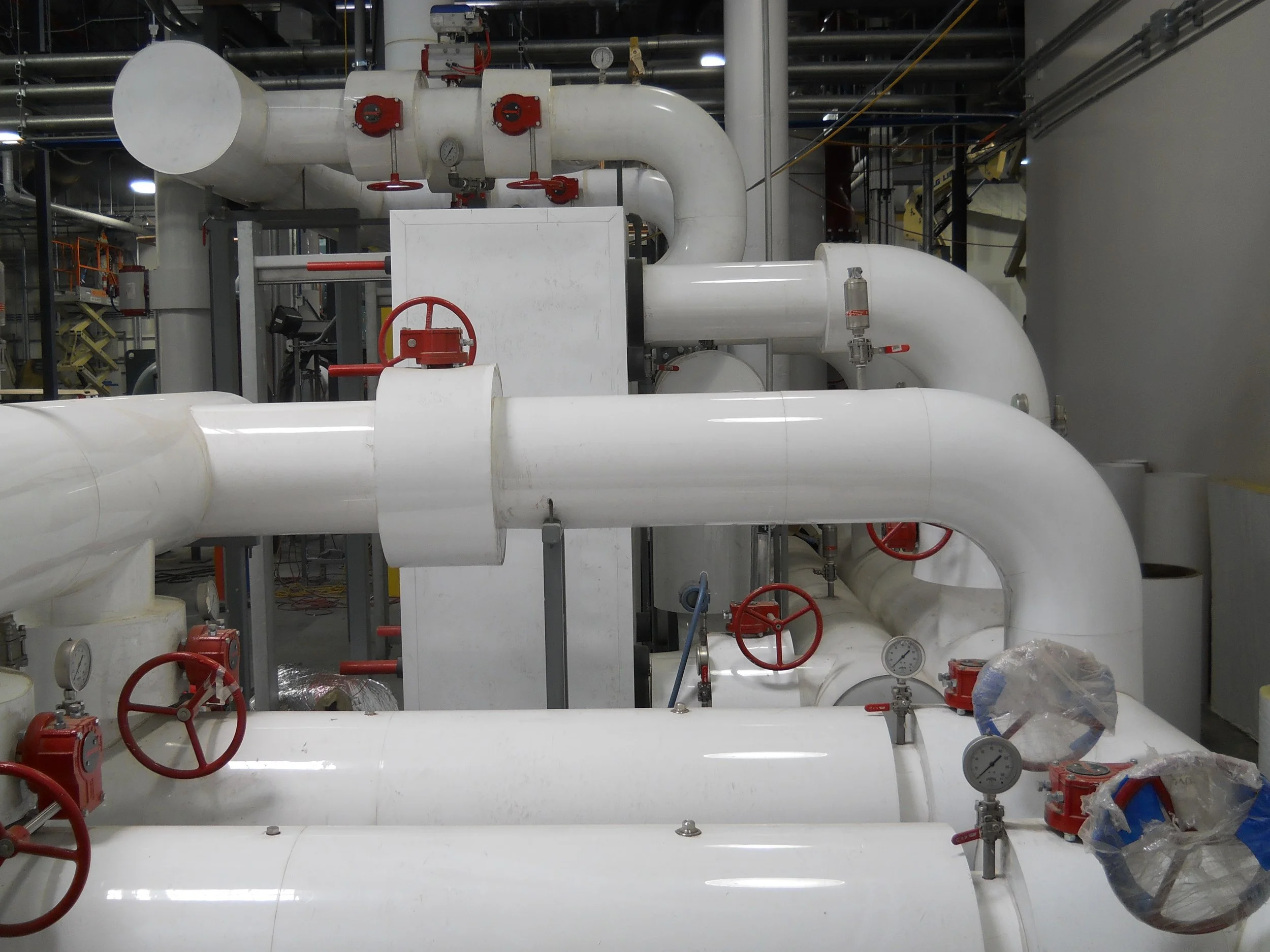 Large white industrial pipes with red valves and pressure gauges in a factory or plant setting, with metal scaffolding and equipment in the background.