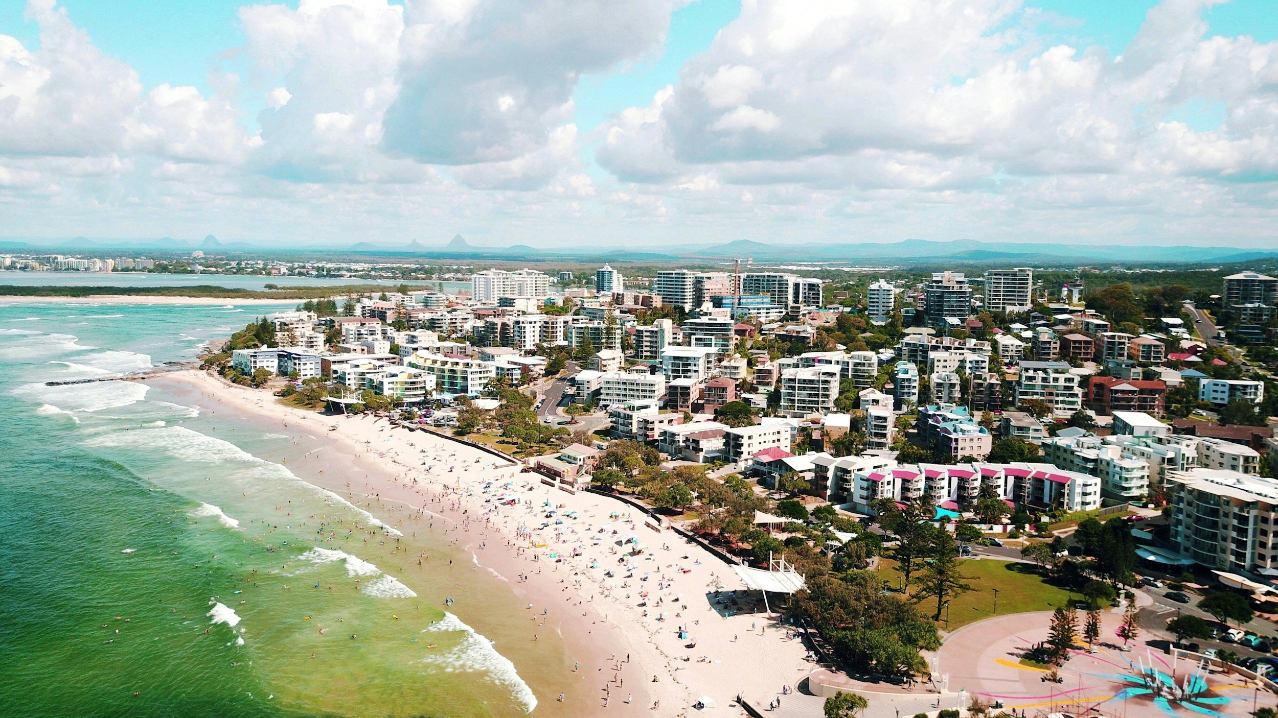 Aerial view of a coastal city with a beach, high-rise buildings, and a cloudy sky.