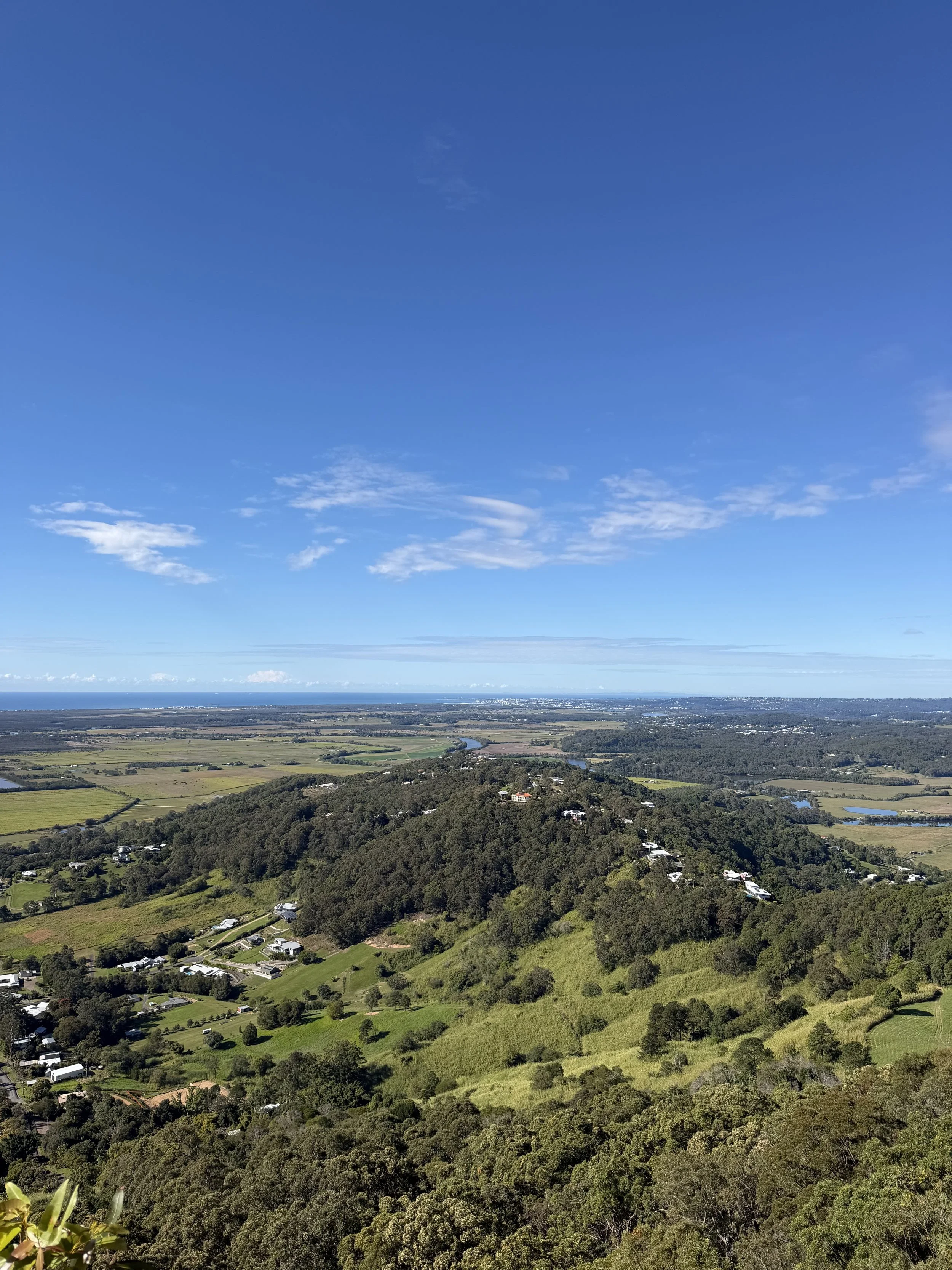 Aerial view of lush green hills, farmland, a river, and scattered houses under a bright blue sky with some clouds.