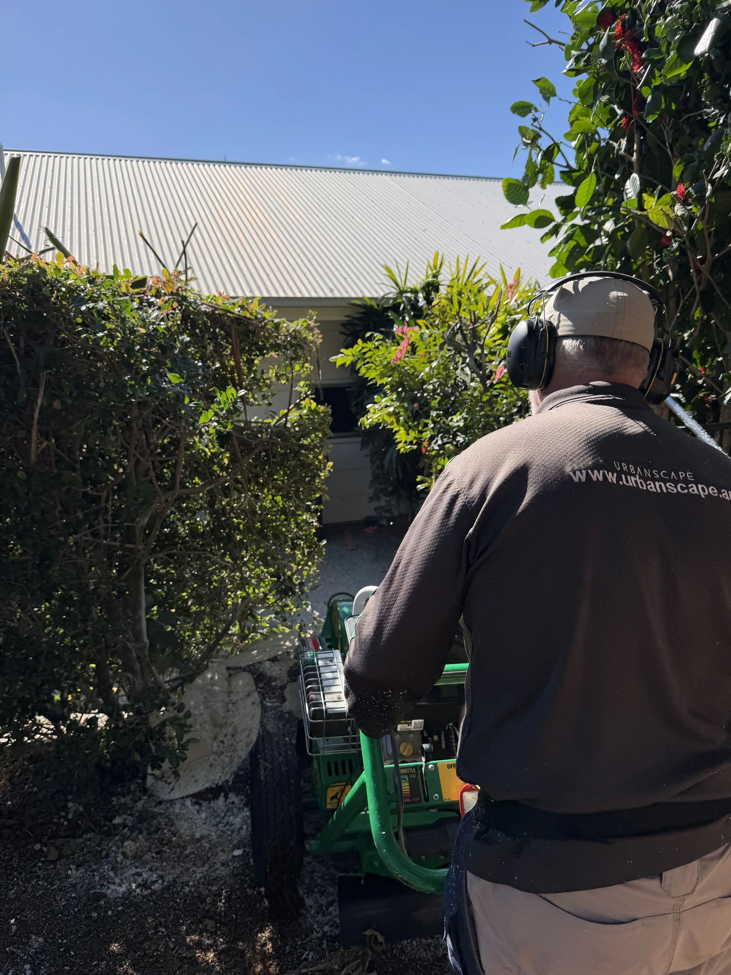 A person wearing a beige cap and dark gray shirt with headphones operating a green machine outdoors, surrounded by bushes and a house with a metal roof under a clear blue sky.
