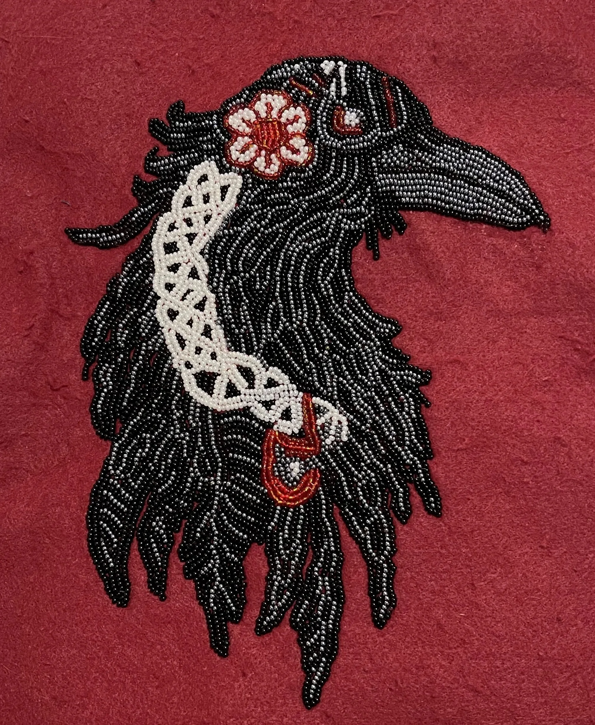 Beaded artwork of a black horse head with a floral design near the ear, on a red background.