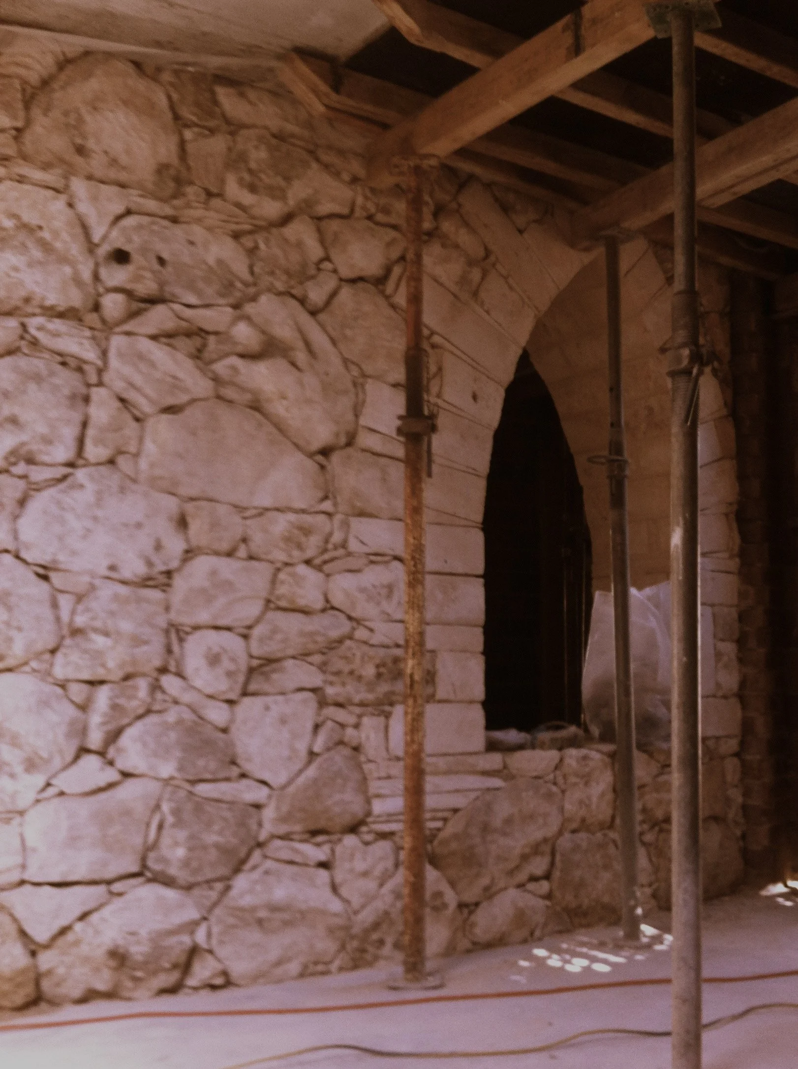 The image shows an interior stone wall with an archway, supported by a large steel pole, and part of the ceiling made of wooden beams.