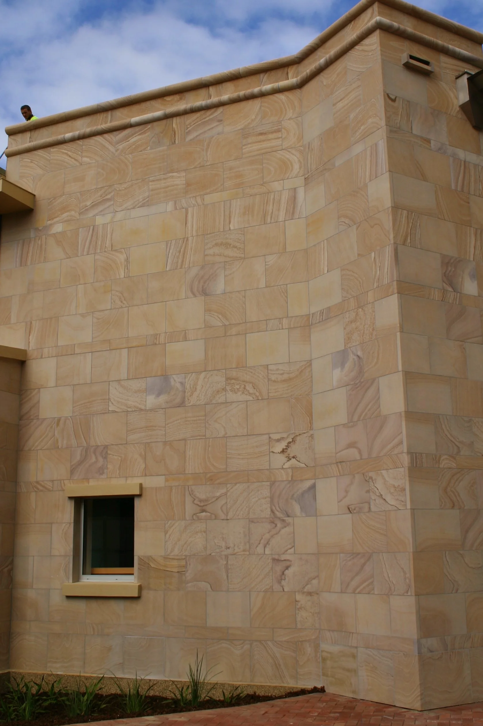 Close-up of a tan-colored stone building with a small window, clouds in the sky, and some plants at the bottom.