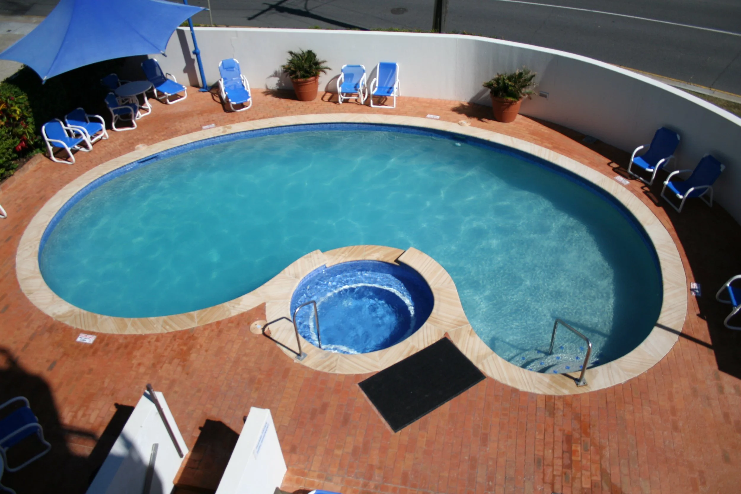 An aerial view of a kidney-shaped swimming pool surrounded by a brick patio, with blue lounge chairs, a large blue umbrella, potted plants, and a white perimeter wall.