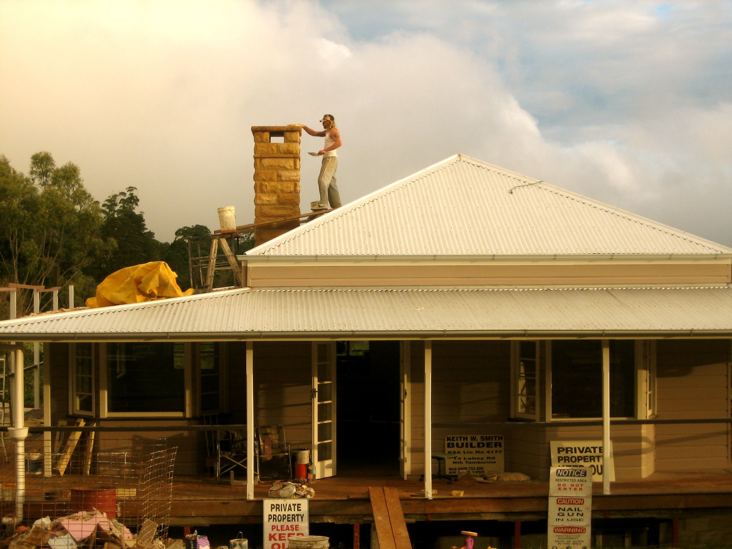 A construction worker standing on the roof of a house installing a chimney. The worker is wearing a cap and sunglasses, holding tools, and working on the chimney. The house has a white corrugated metal roof, and there are trees and a cloudy sky in the background.