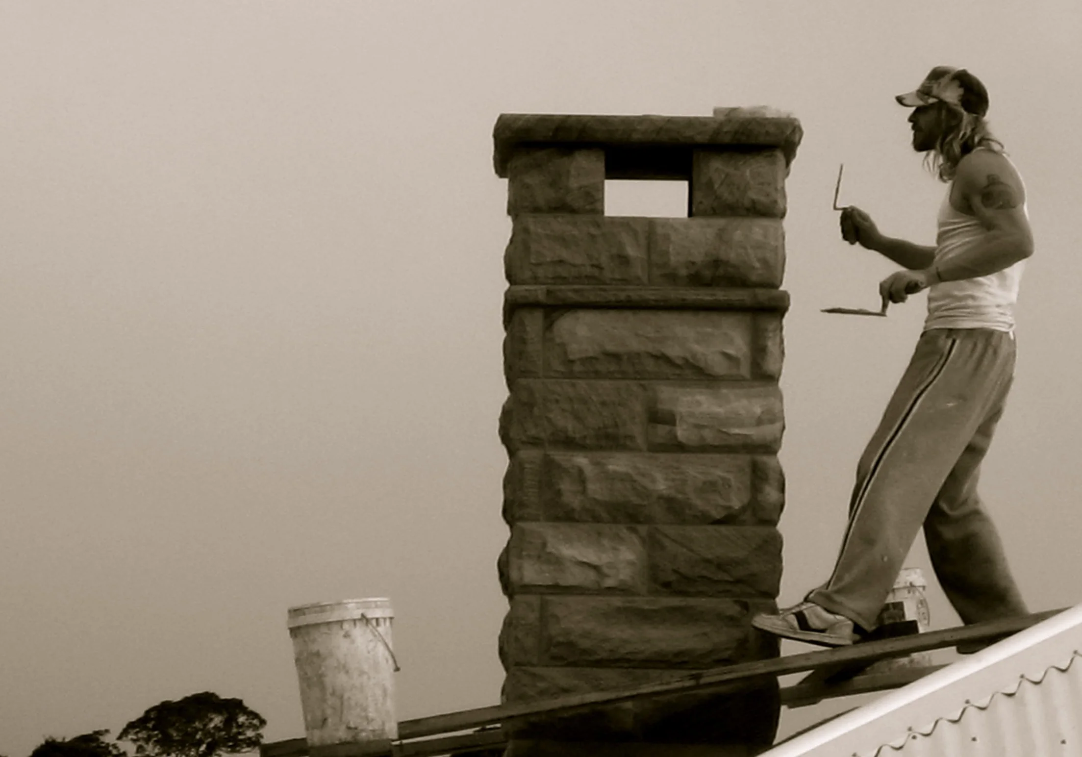 A person working on a chimney made of large stone blocks, wearing a cap, sleeveless shirt, sweatpants, and sneakers, standing on a platform, with a bucket and tools nearby.