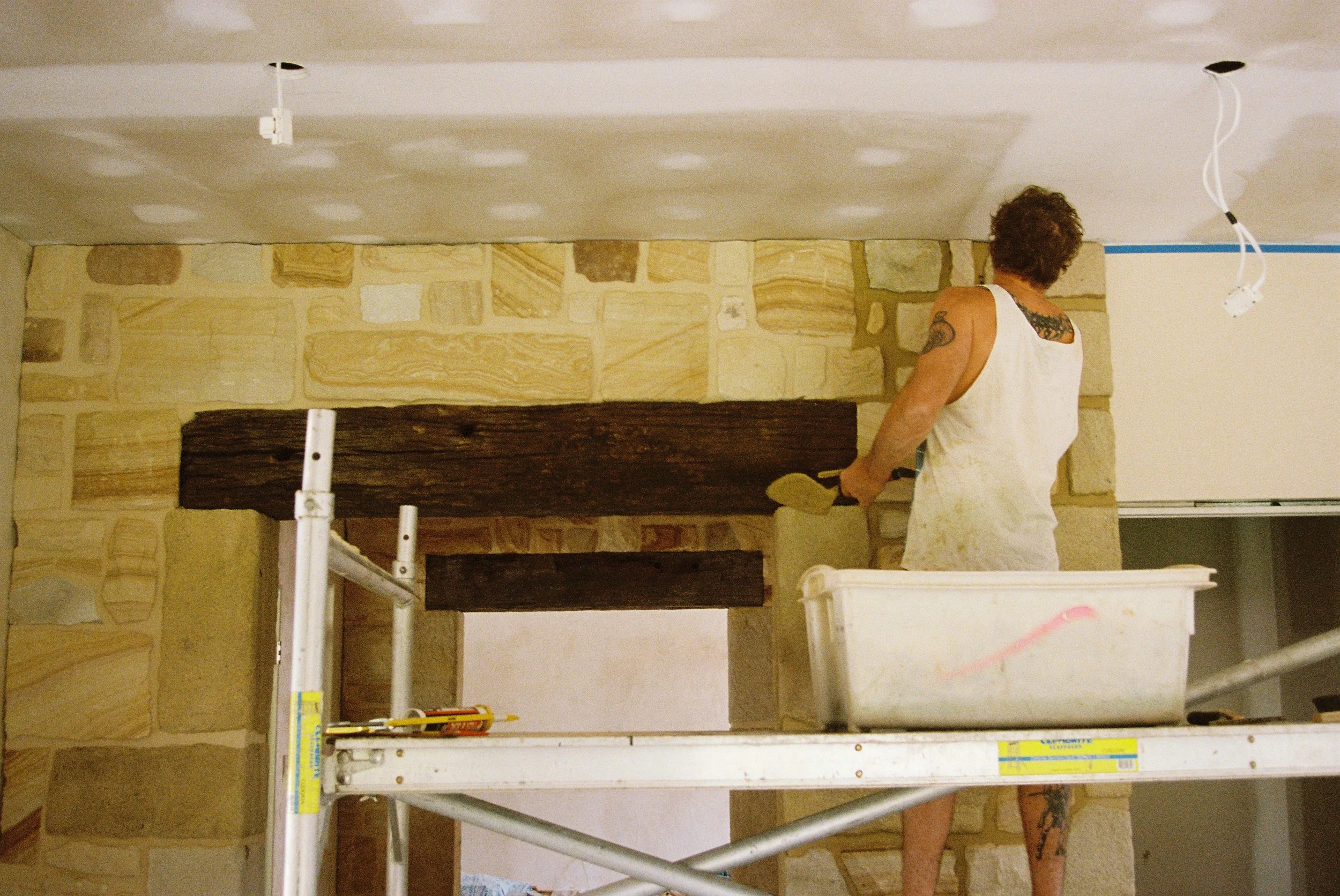 A person working on a ceiling, with part of their body and tattoos visible, standing near scaffolding, in a room with stone walls and unfinished ceiling.