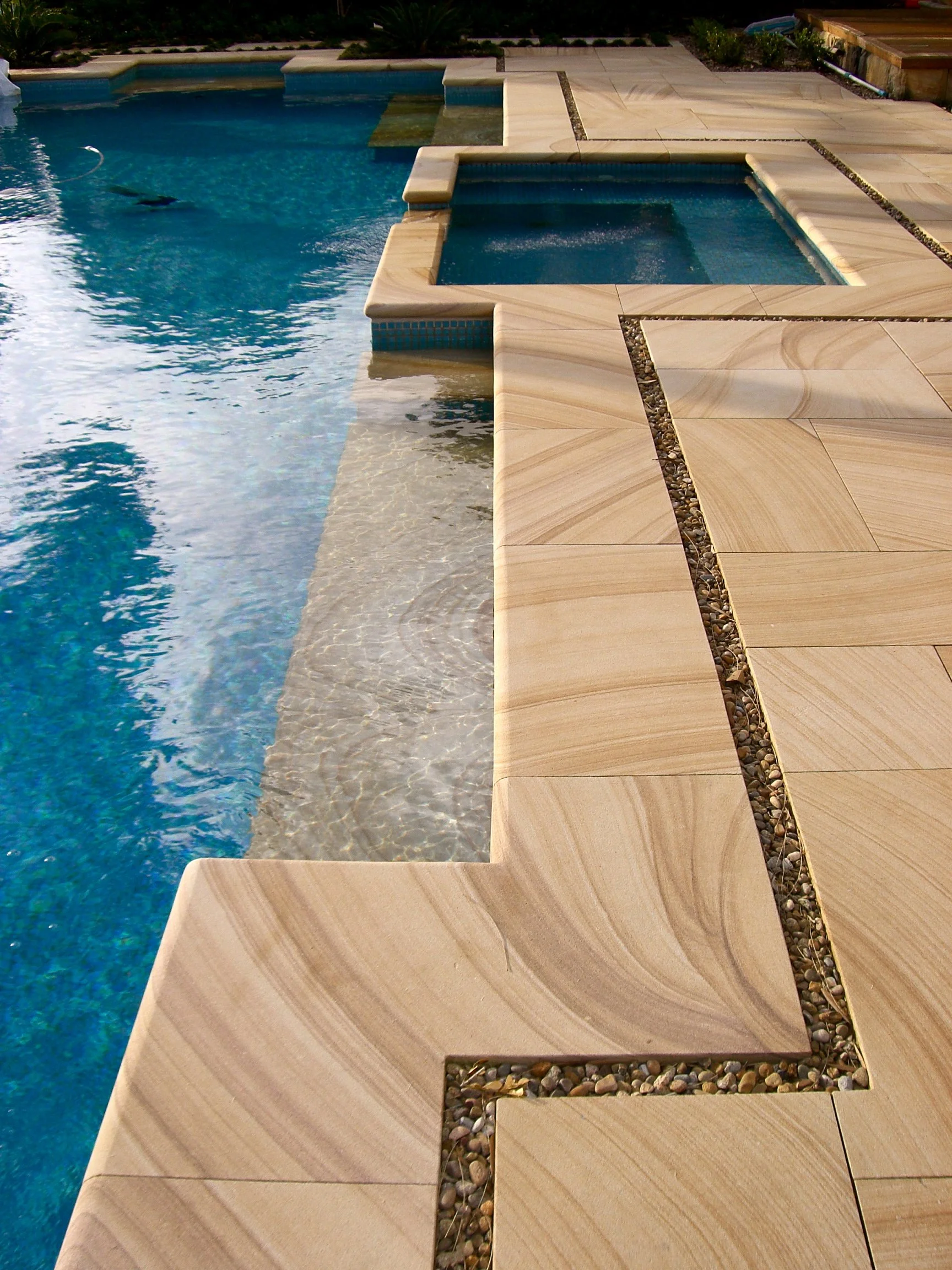 A backyard swimming pool with a submerged spa on a stone patio with small decorative rocks in between the tiles.
