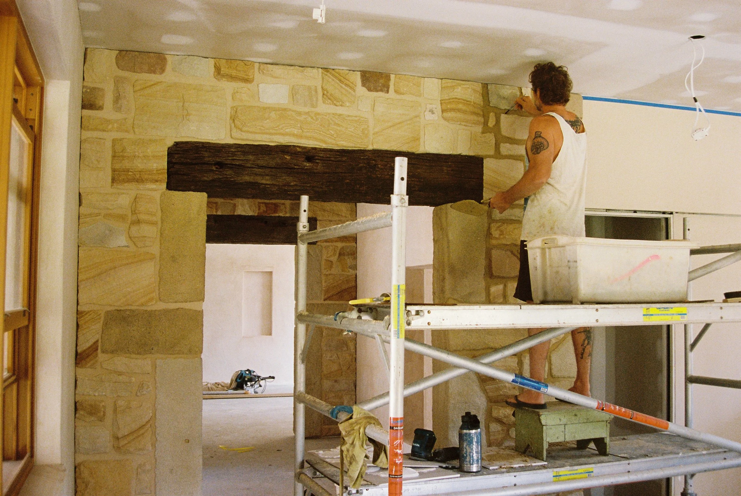 A worker standing on scaffolding painting a room's wall, which has stone veneer partially installed. The worker is holding a paintbrush and wearing a sleeveless shirt and shorts.