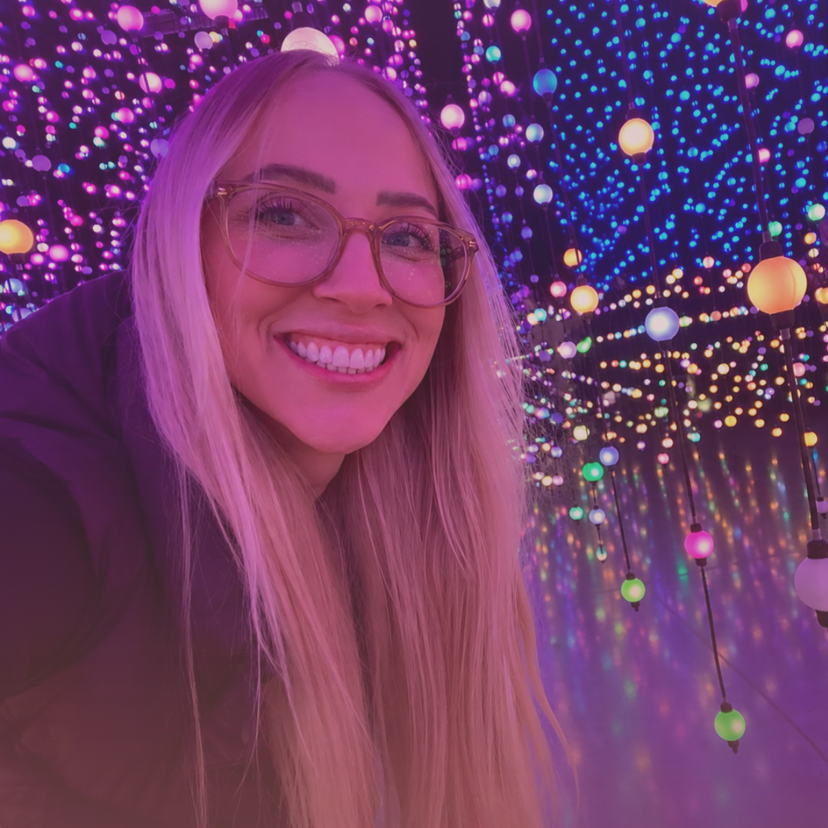 A smiling woman with blonde hair and glasses taking a selfie in an immersive LED light art installation with hanging colorful LED orbs and reflections.