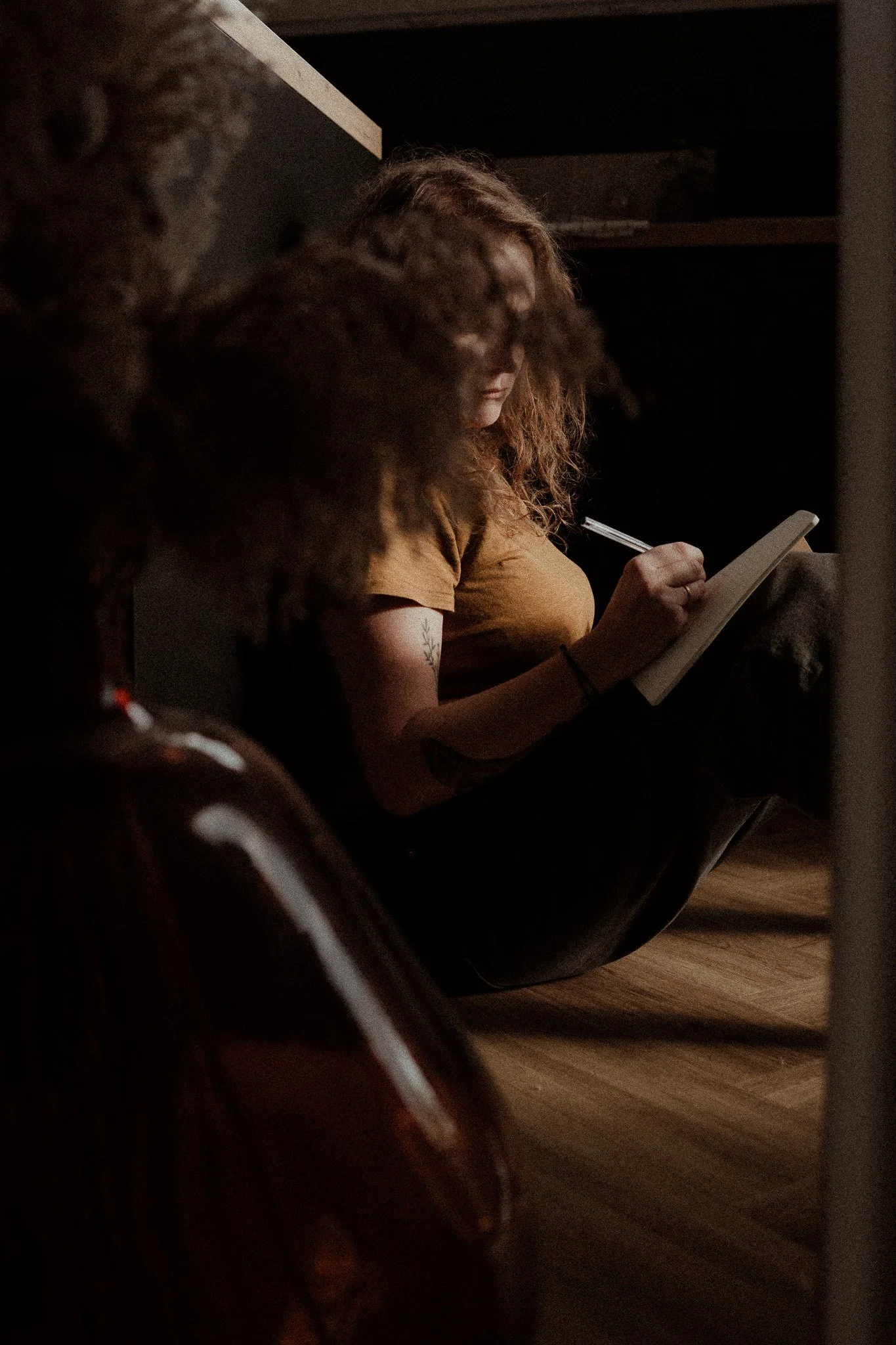 Une femme aux cheveux bouclés, portant un t-shirt marron, lit ou écrit dans un carnet, dans un environnement sombre pour poser les mots et les maux lors d'un accompagnement personnalisé pour les femmes en Alsace près de Colmar.