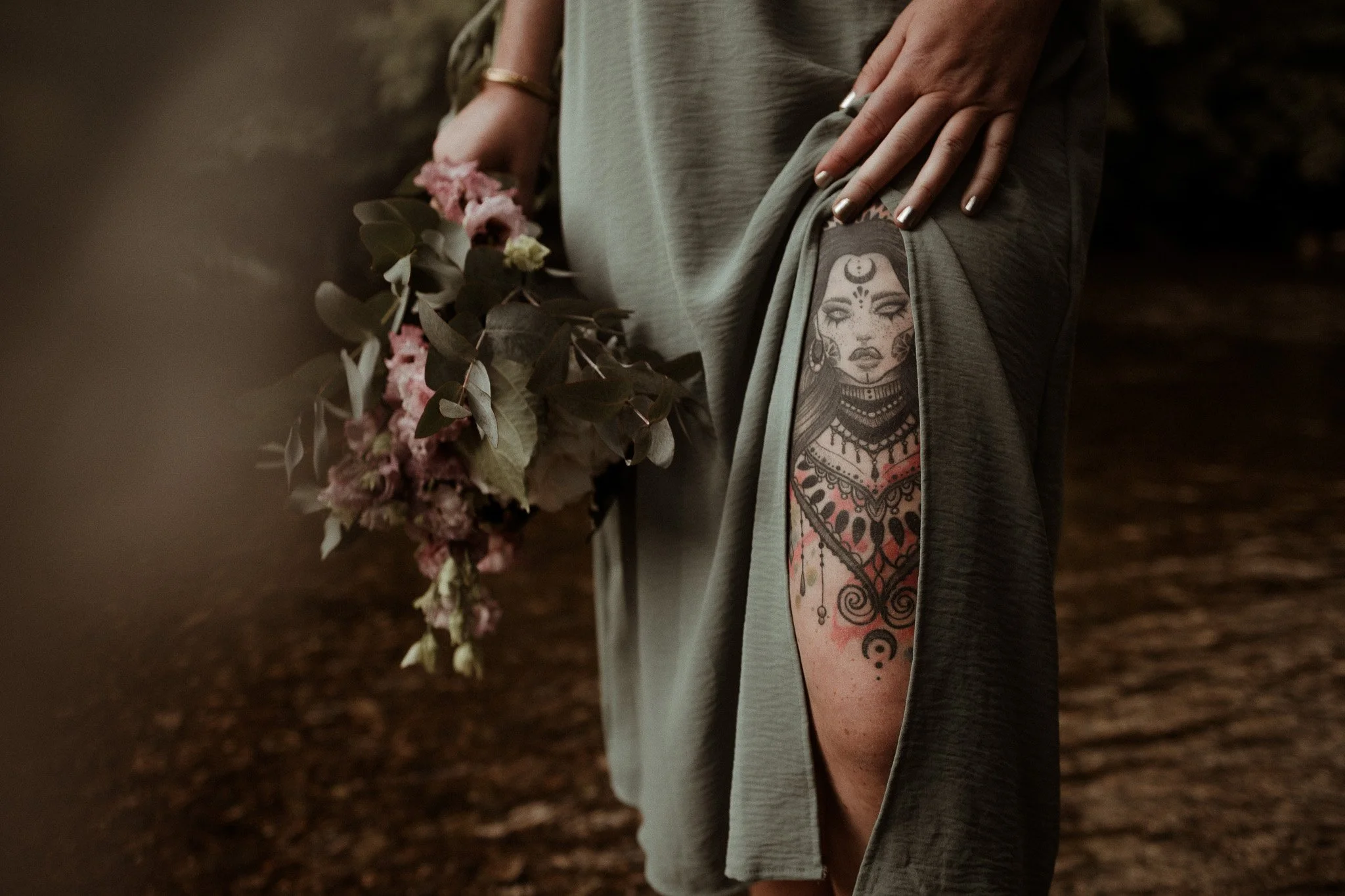 Close-up d'une femme s'offrant une séance photo pour elle, un bouquet de fleurs, portant un débardeur gris, avec un tatouage coloré sur le bras. La personne porte également une bague et un bracelet, et se tient dans une zone sombre ou boisée.