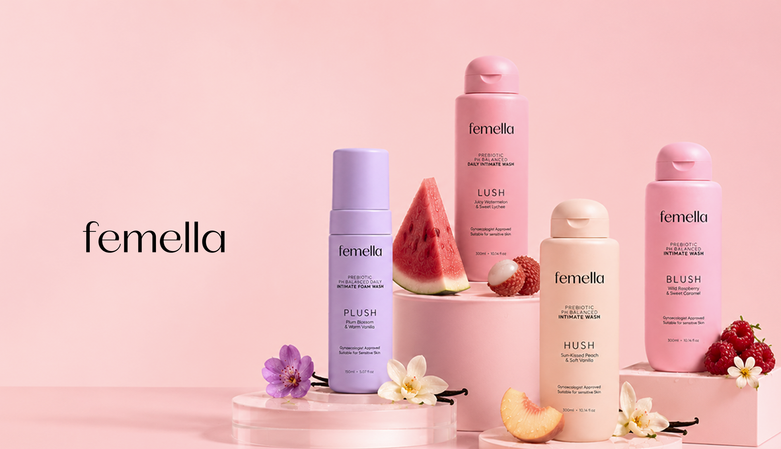 Arrangement of Femella skincare products including foams and washes in pink, purple, and beige bottles, with watermelon, raspberries, flower petals, and citrus slices on a pink background.