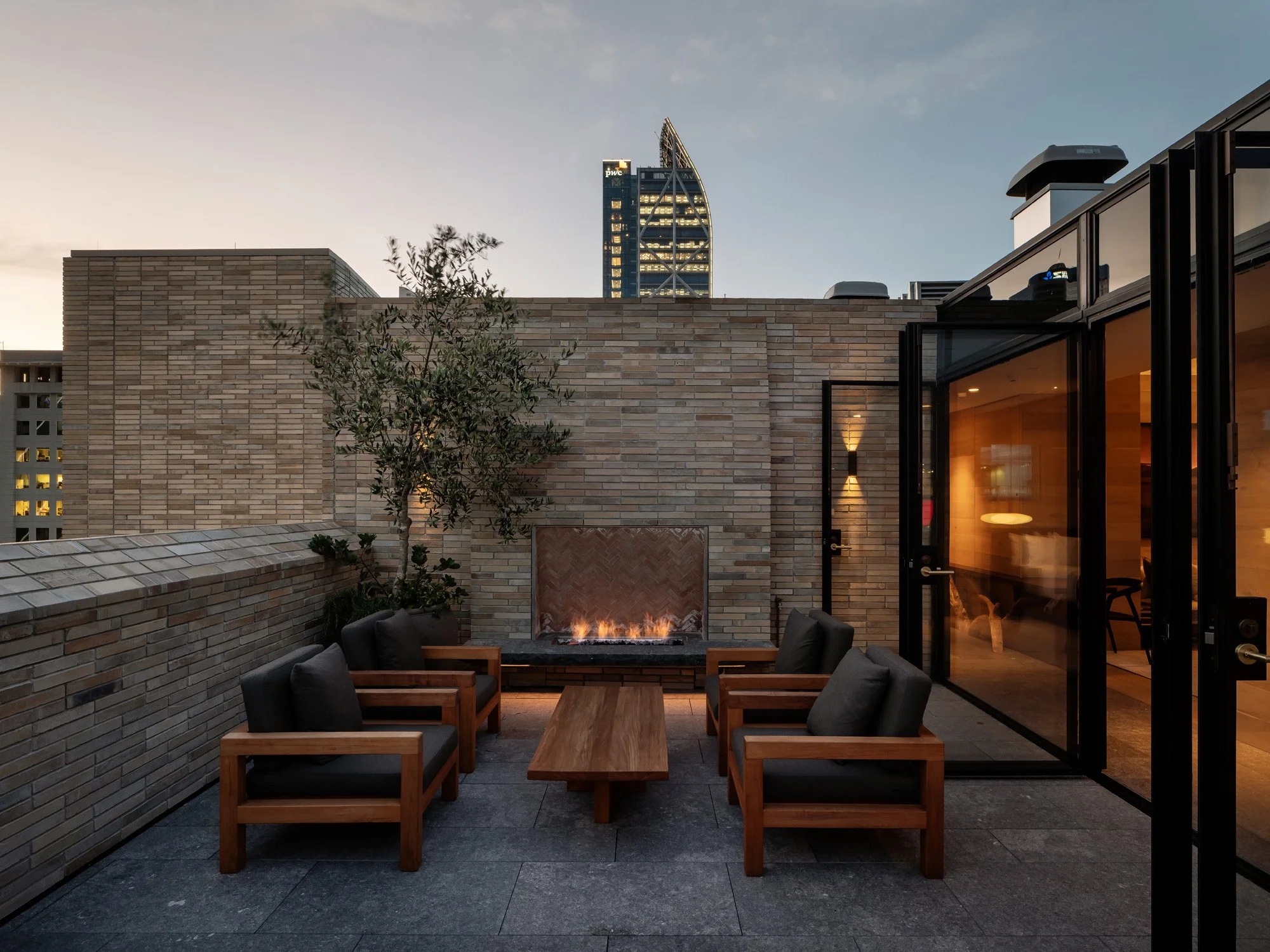 Urban rooftop patio with brick walls, outdoor fire pit, and modern seating, overlooking city skyscrapers at dusk.