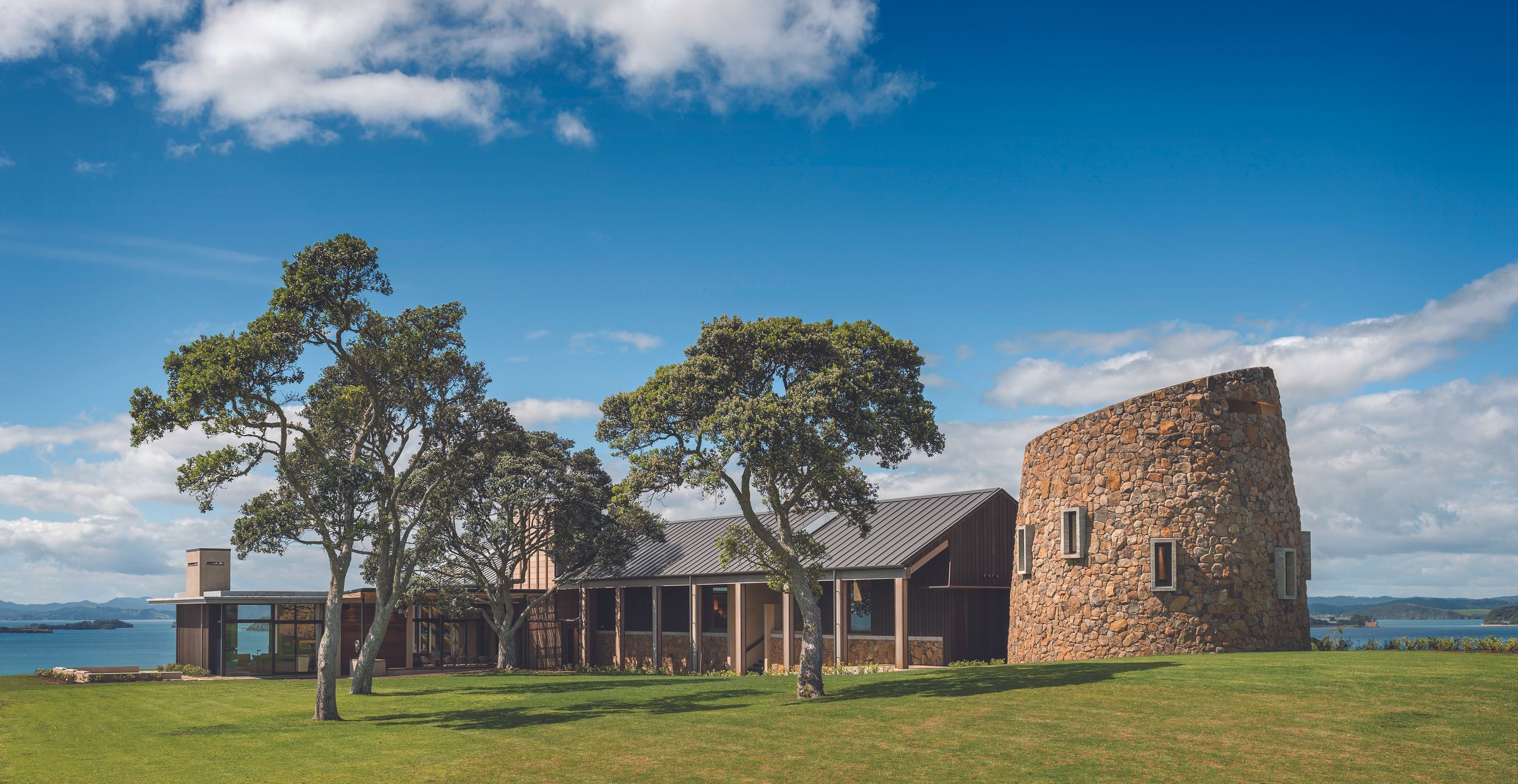 A modern house with a curved stone tower, large windows, and a metal roof, surrounded by trees and a grassy lawn near water under a cloudy blue sky.