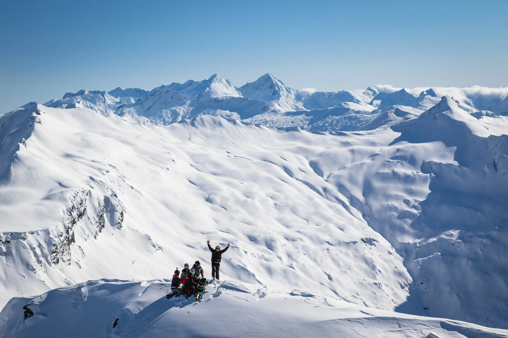 A group of people in winter gear on snowy mountain terrain at high altitude, with a person standing on a rock raising their arms, and snow-capped mountains in the background under a clear blue sky.