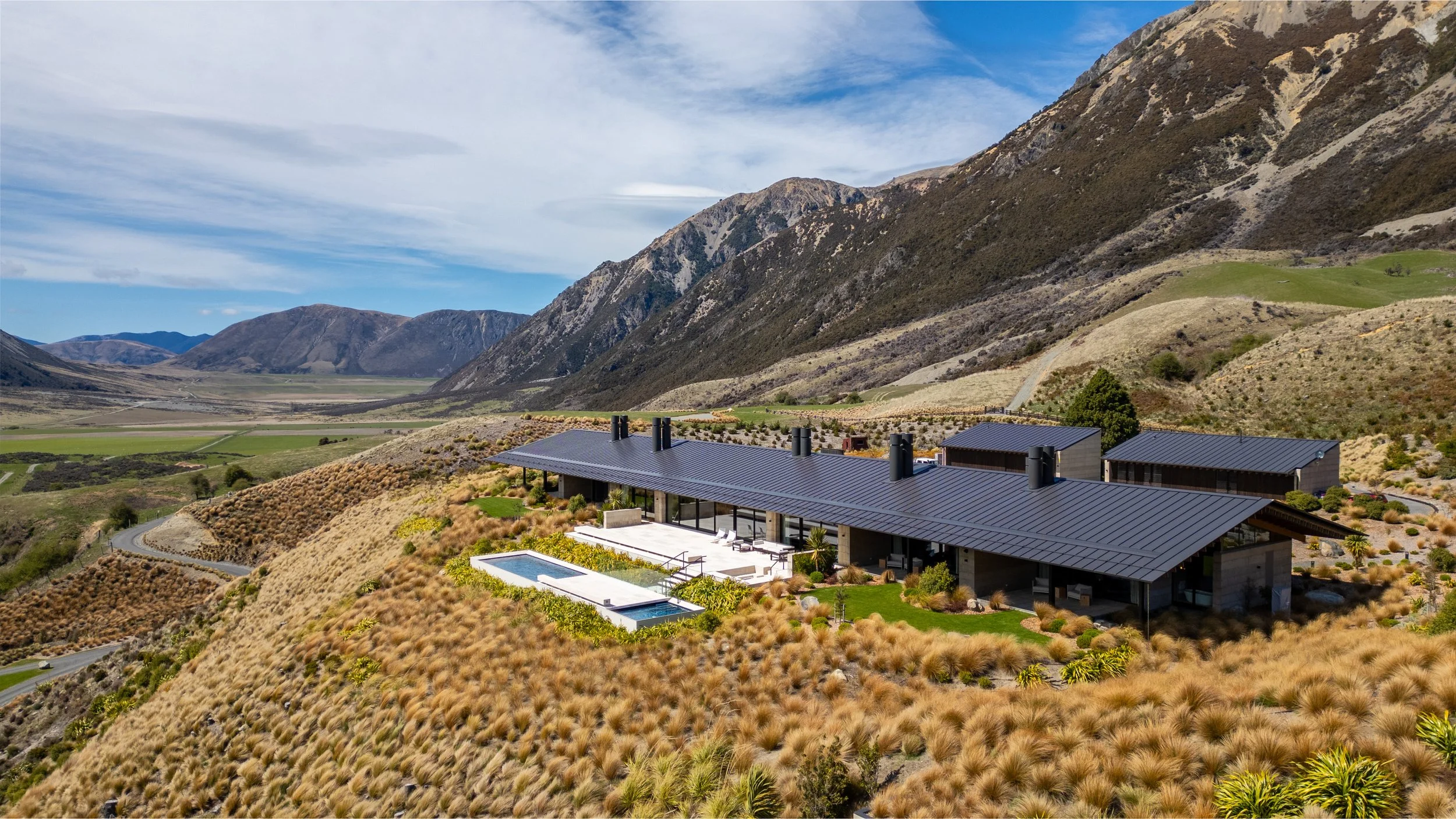 Modern house with black metal roof and outdoor pool, situated on a hillside with brown and green vegetation, overlooking a valley and mountains under a partly cloudy sky.