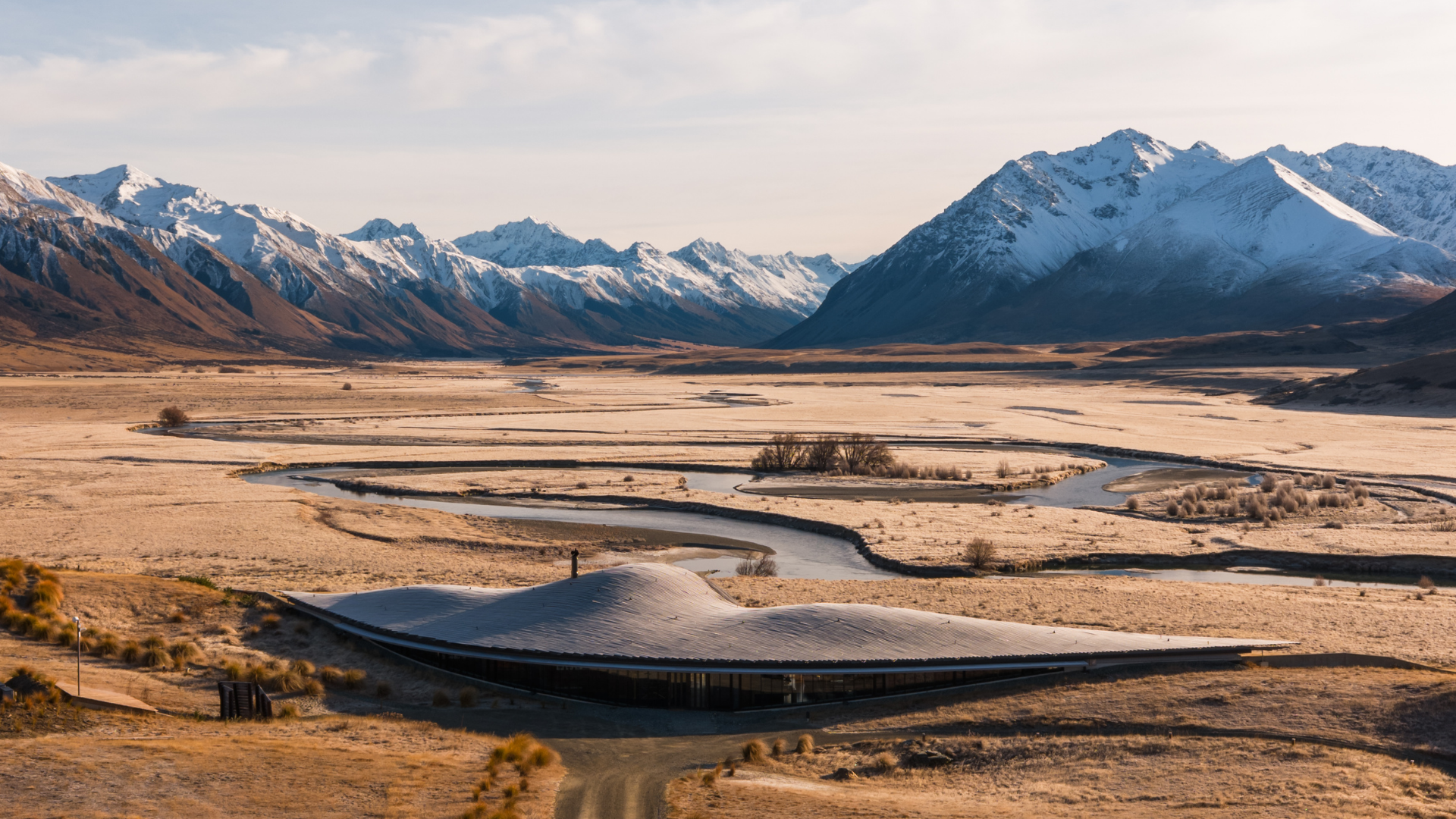 Scenic landscape with winding river, snow-capped mountains, and a modern curved building in the foreground.