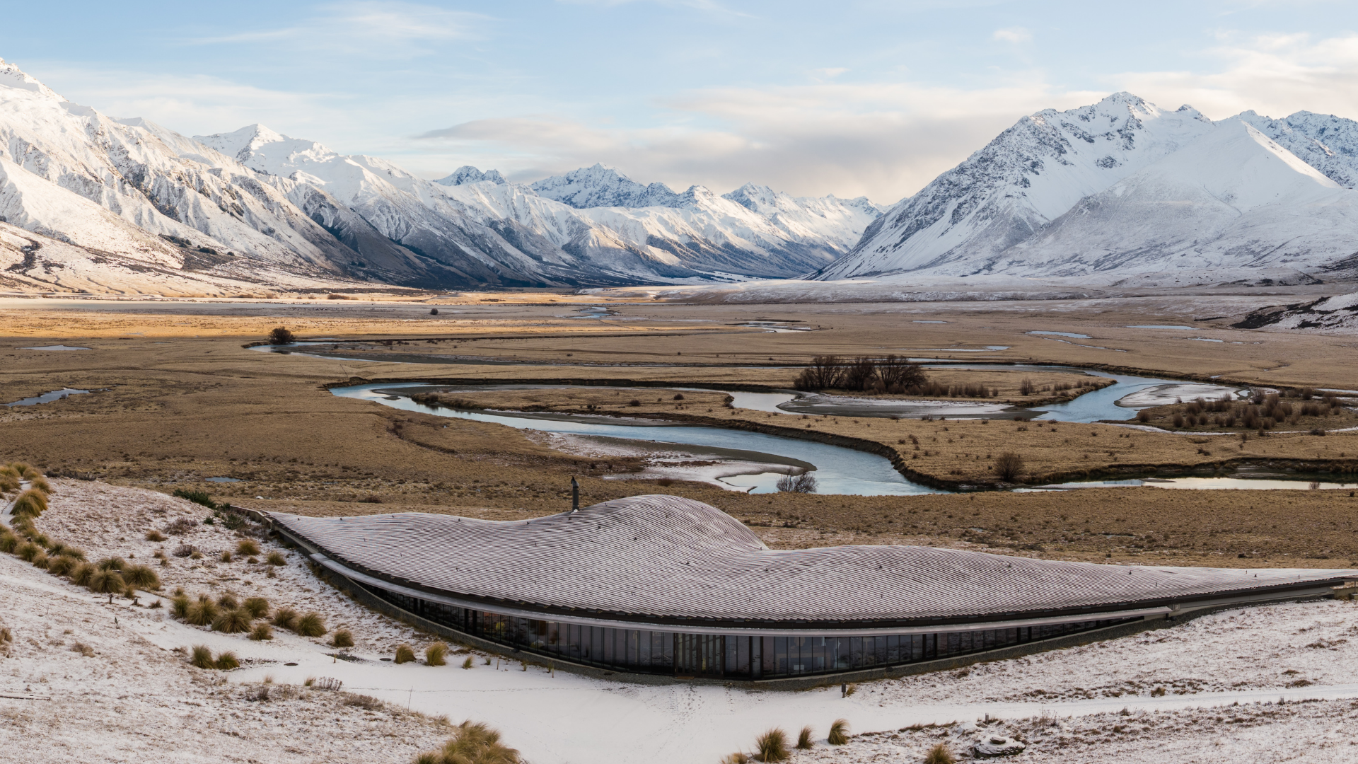 Modern building with curved roof in snowy landscape with mountains and winding river in background.