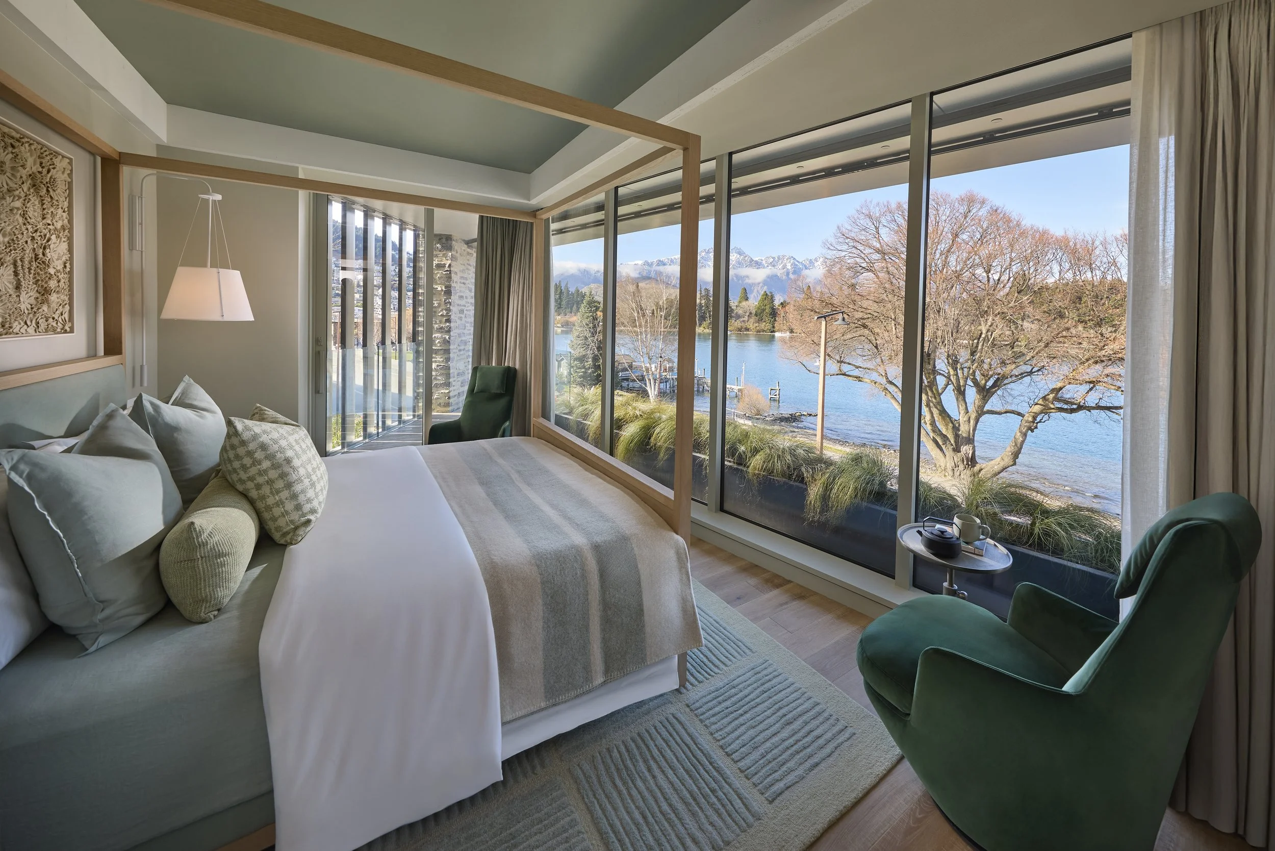 A bedroom with large glass windows offering a view of a lake, trees, and snow-capped mountains in the distance. The room has a bed with light-colored bedding and pillows, a green armchair, a small round table with a cup and a book, and a hanging light fixture.