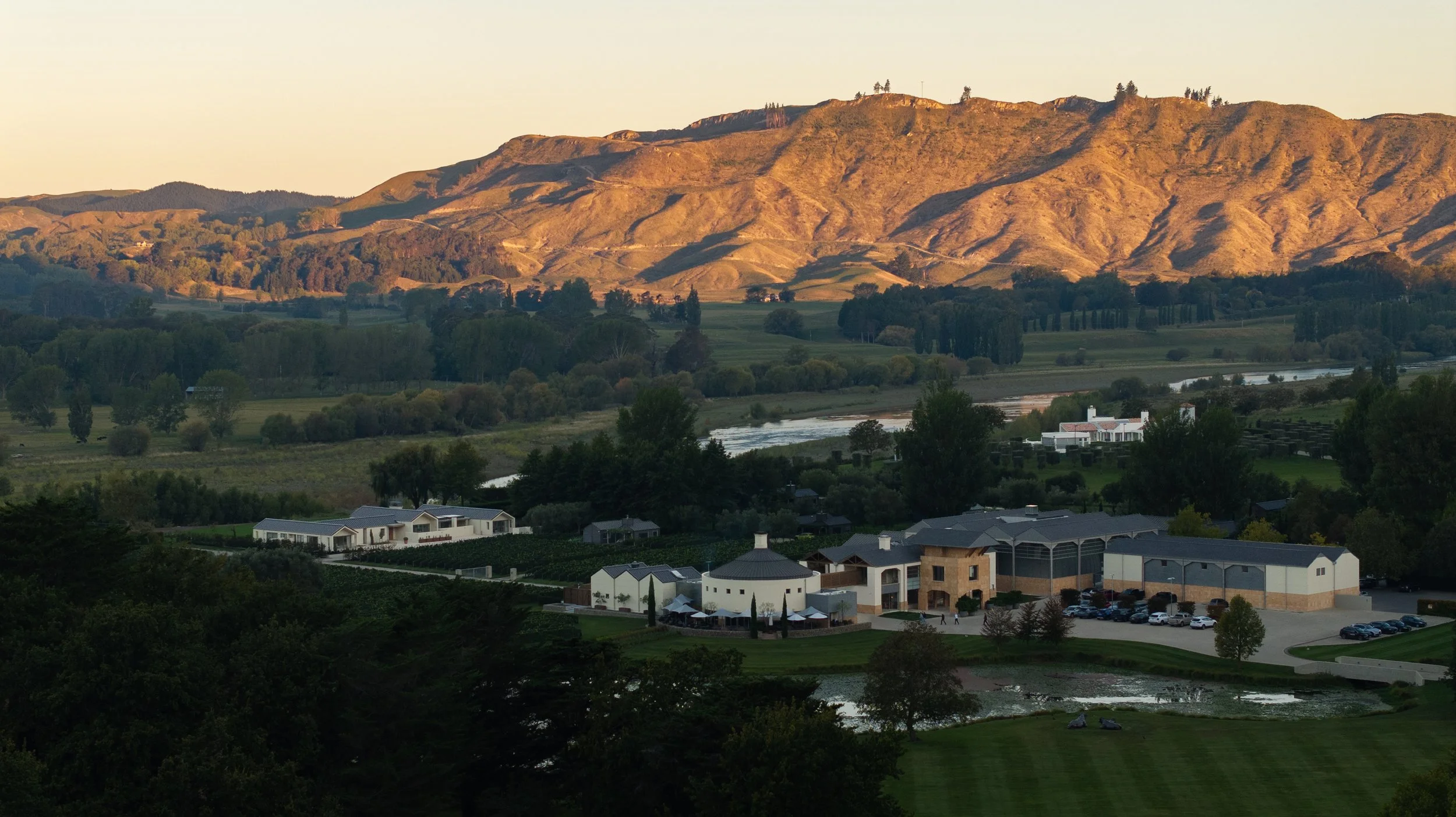 Scenic landscape with rolling hills, trees, a river, and buildings, likely in a suburban area at sunset.