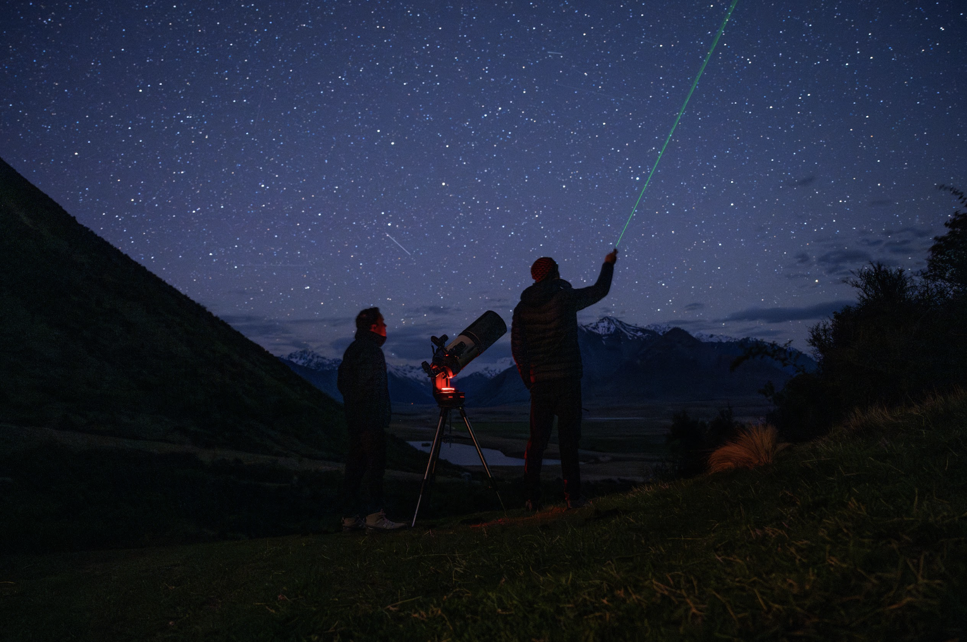 Two people stargazing at night with a telescope, one of them pointing a green laser into the starry sky, mountainous landscape in the background.