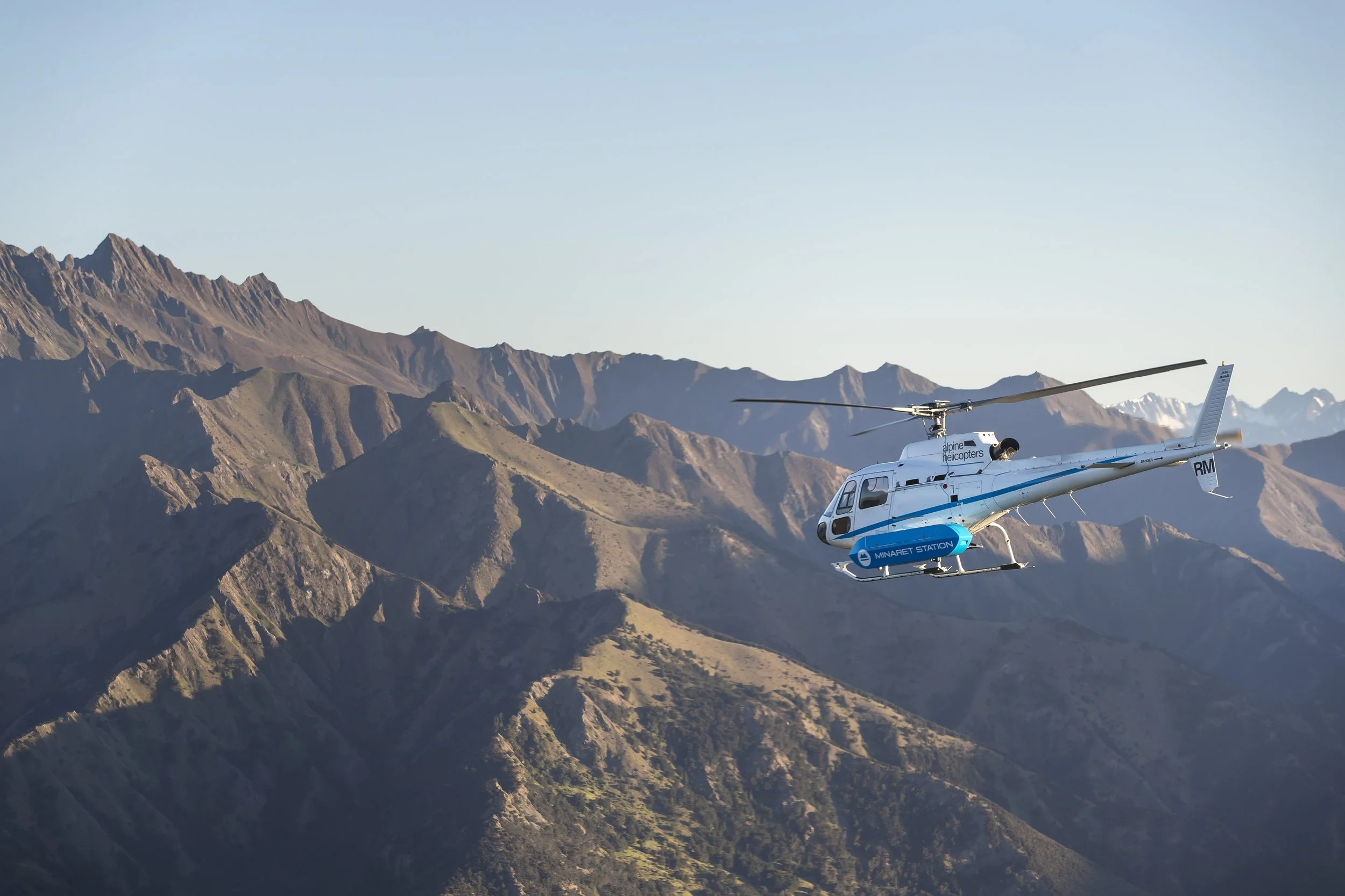 A white helicopter flying over mountainous terrain with rugged, rocky peaks under a clear sky.