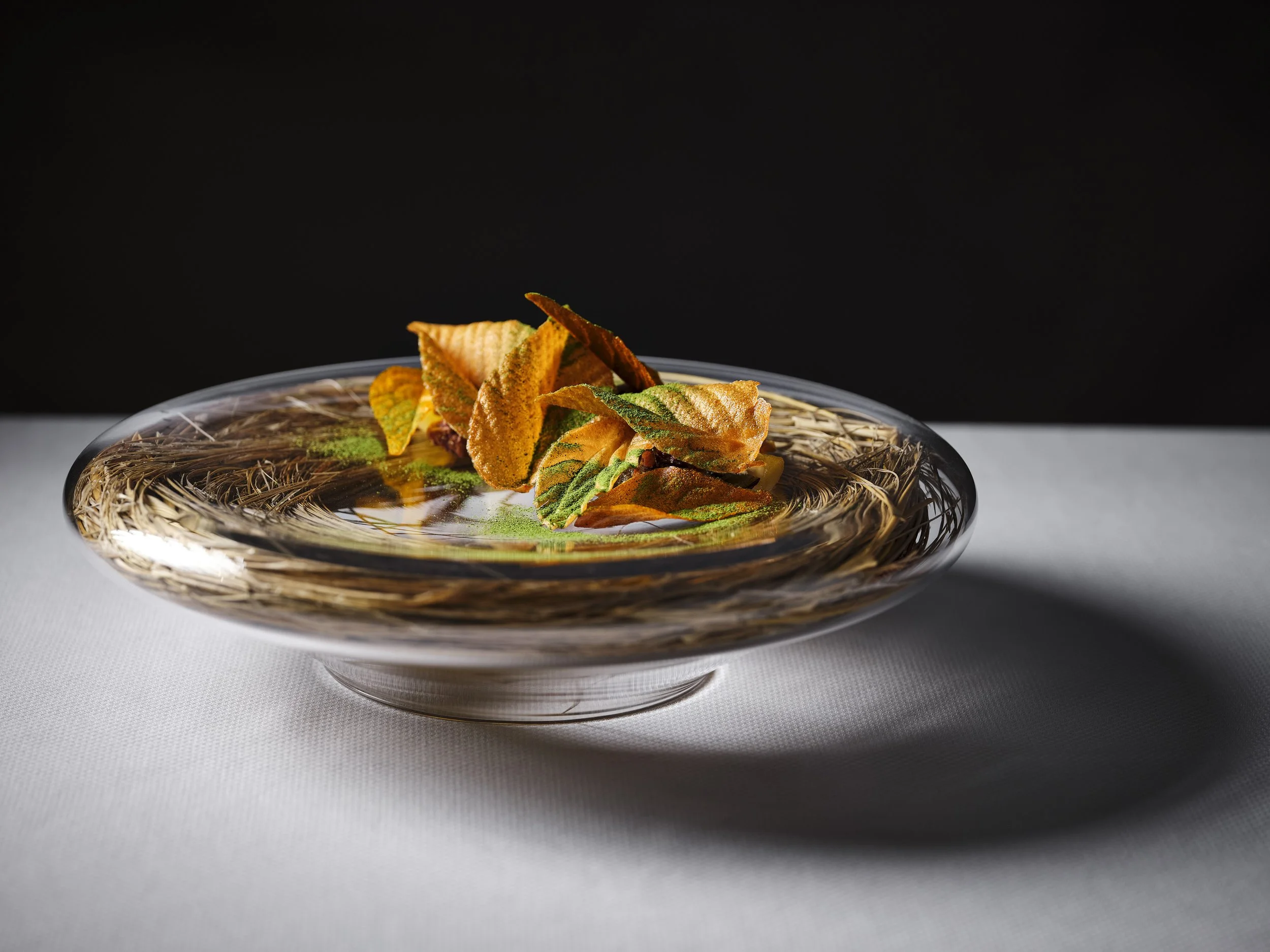 A clear glass bowl with intricate design containing fried chips and green herbs on a white surface with a black background.