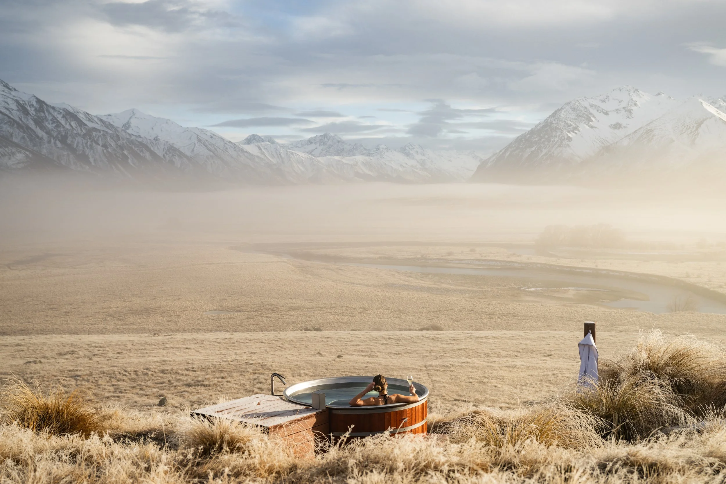 A woman relaxing in a hot tub outdoor on a grassy area with mountains in the background, during a cloudy day.