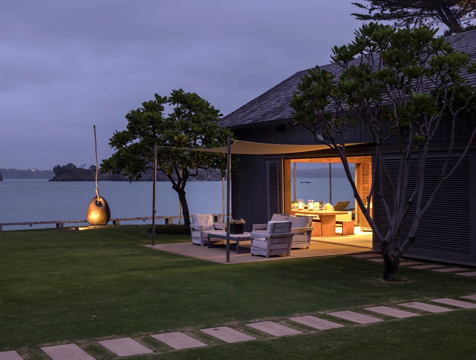 A beachside patio with outdoor seating, trees, a decorative lantern, and a view of the ocean during dusk.