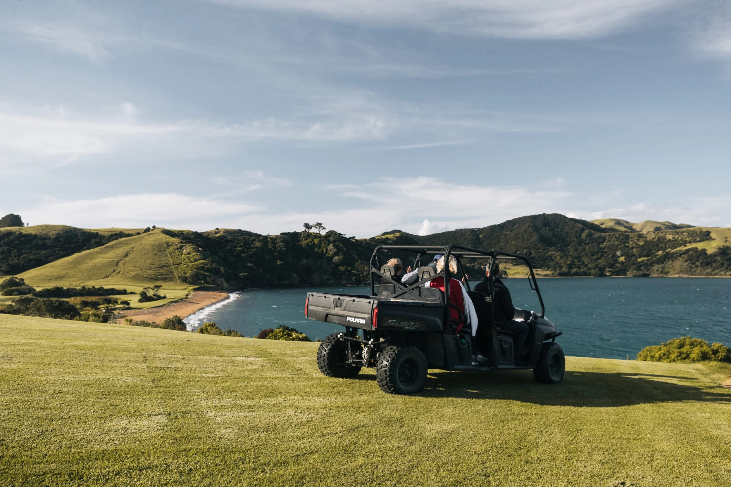 Off-road utility terrain vehicle (UTV) parked on grass overlooking a body of water, with rolling hills and a blue sky with clouds in the background, and three people seated inside the vehicle.