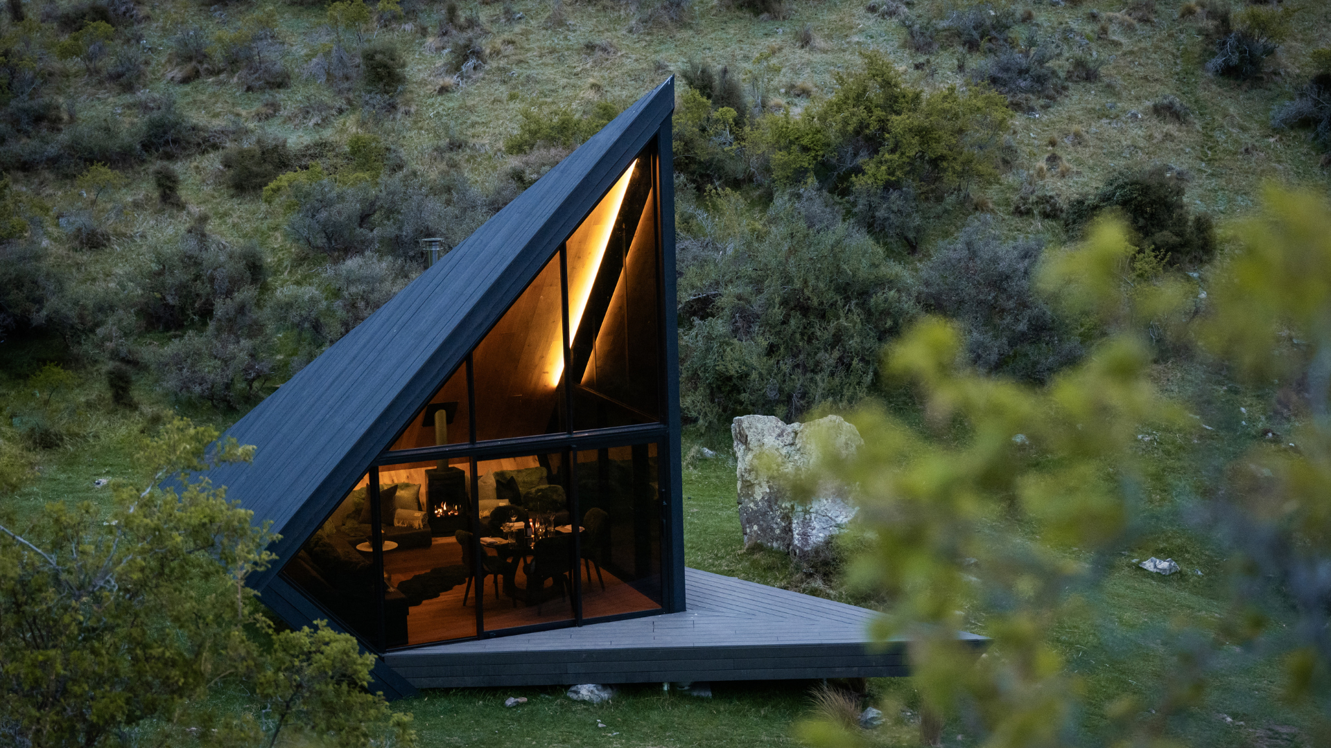 A modern house with a steep, triangular roof, illuminated interior visible through large glass windows, situated in a green, hilly landscape with trees and rocks, during dusk.