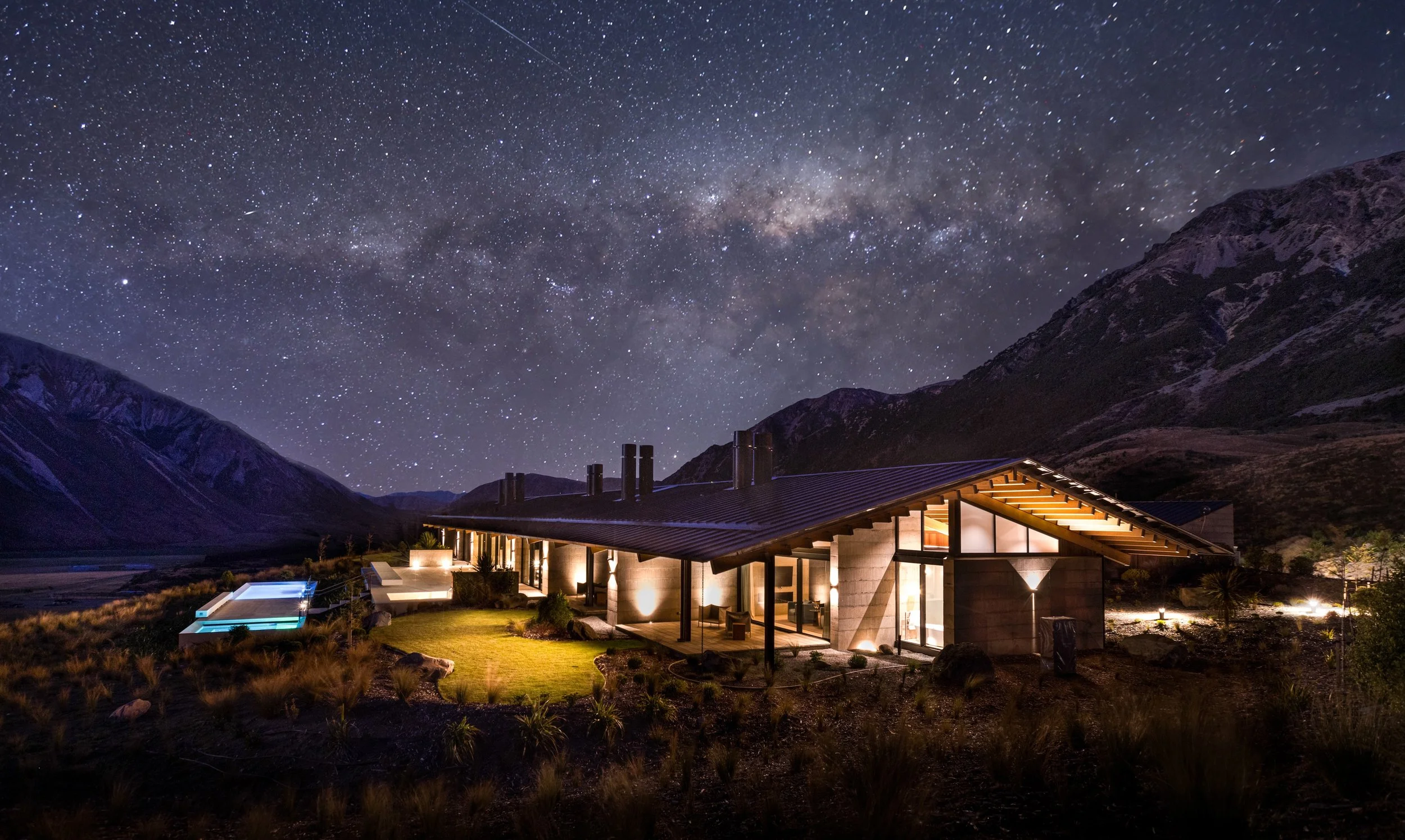 A modern house with large windows in a mountainous landscape at night, illuminated by outdoor lighting, under a star-filled sky with the Milky Way galaxy clearly visible.