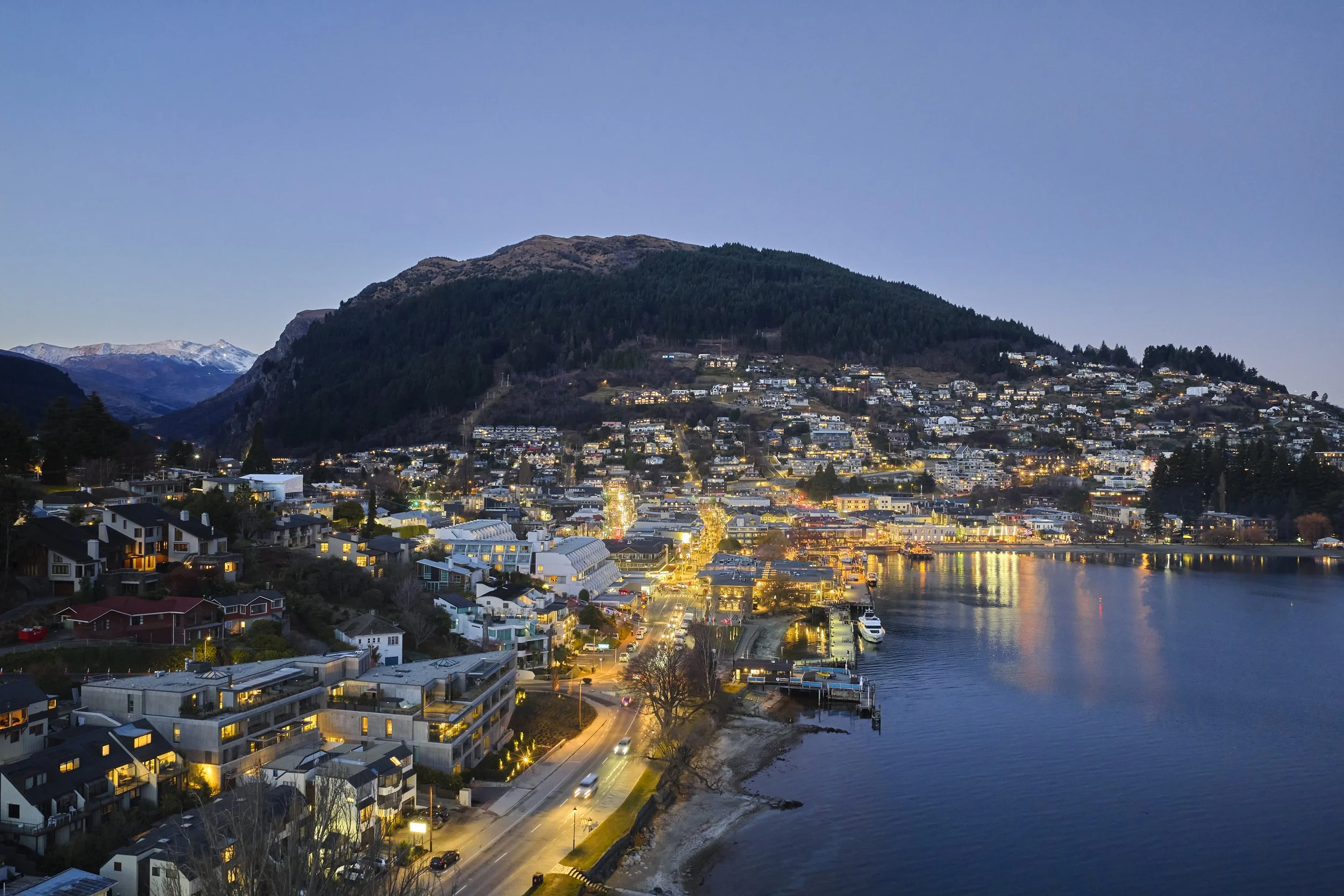 Nighttime cityscape with hillside residential area, a body of water reflecting streetlights, and snow-capped mountains in the background.