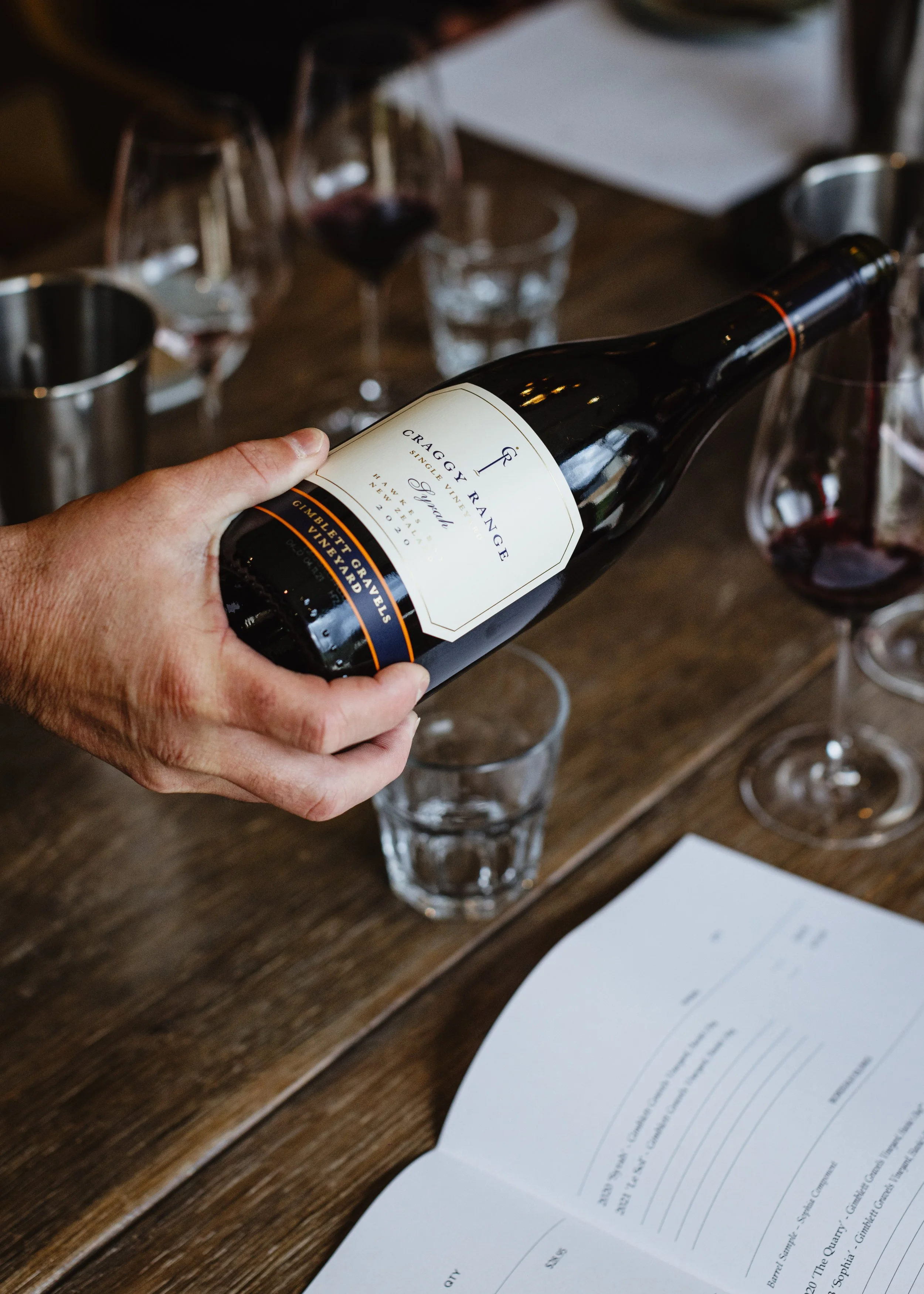 Person holding a bottle of wine labeled Craggy Range, surrounded by glasses, a notepad, and a pen on a wooden table.