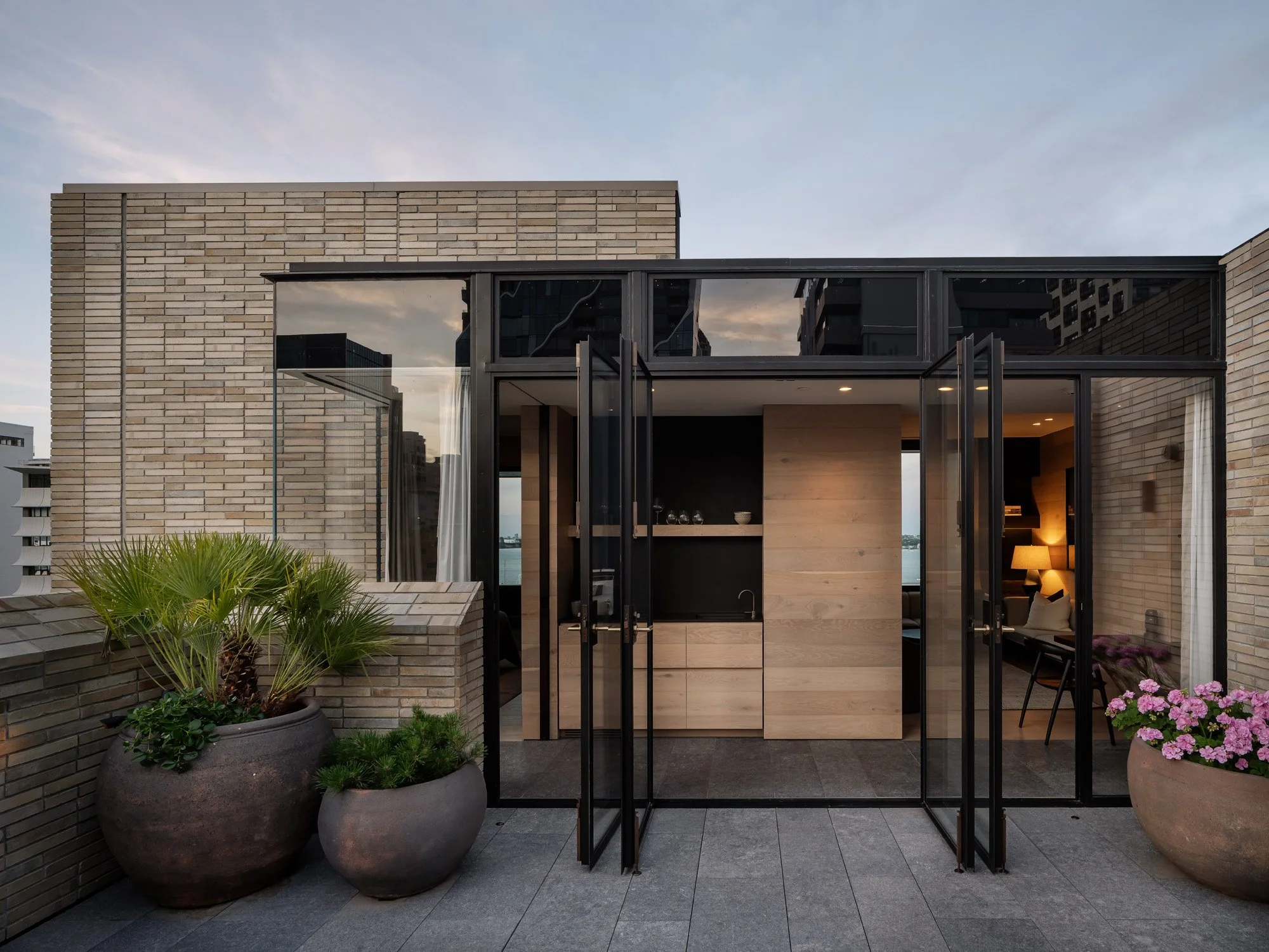 Modern rooftop terrace with large potted plants, brick walls, and a glass door leading inside a contemporary apartment
